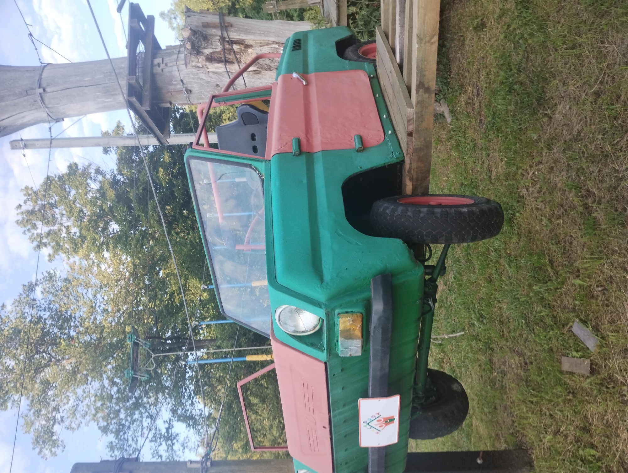 The image shows a modified, green and pink, open-top car, possibly a vintage Soviet-era vehicle, mounted on a wooden platform. The car has a roll cage, a black bucket seat, and a windshield. It is outdoors, with trees and a blue sky visible in the background. A tall, rough-barked wooden pole with attached wooden platforms and wires is to the right of the car, suggesting an adventure park or a similar recreational area. The ground is covered in grass. A small rectangular sign with a colorful graphic is attached to the front of the car. The scene appears to be in an outdoor setting, possibly a park or a recreational facility, with elements that could suggest a themed attraction.