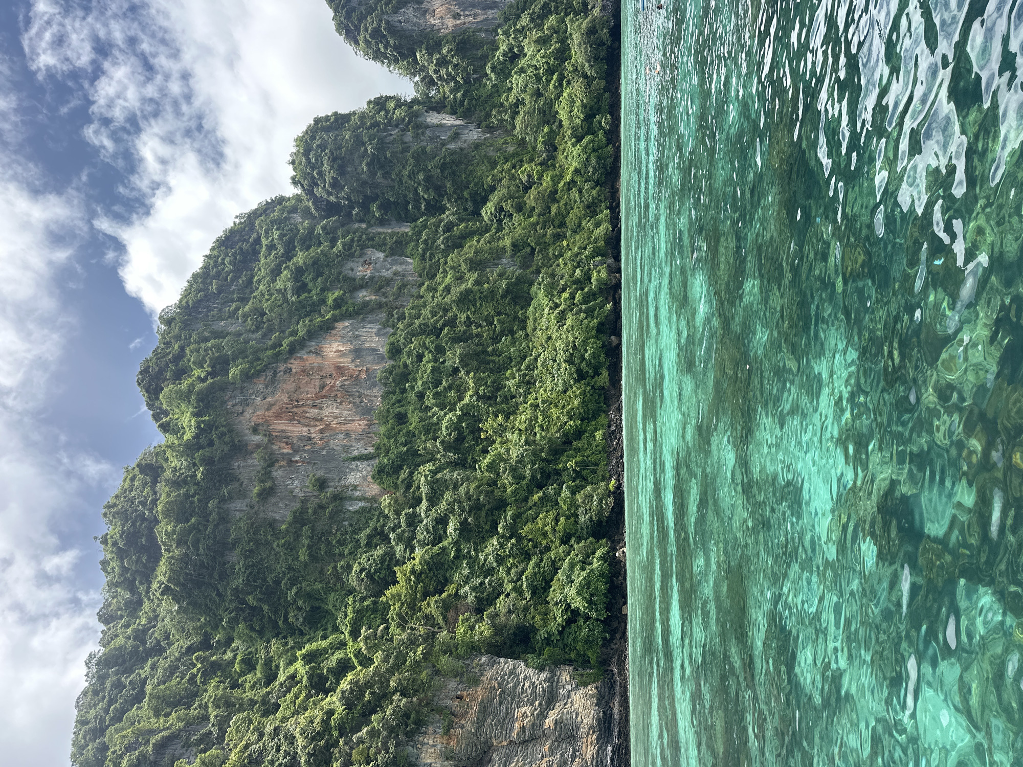 A vertical, eye-level shot captures the vibrant turquoise waters of Ao Nang, Thailand, reflecting the dramatic, verdant limestone cliffs that form the backdrop. The clear water reveals submerged rocks and corals beneath its surface. Lush green foliage blankets the towering cliffs, with patches of bare rock visible where the vegetation has not yet taken hold. The sky above is a mix of blue and white clouds. The scene appears serene and untouched, evoking a sense of natural beauty and tranquility.