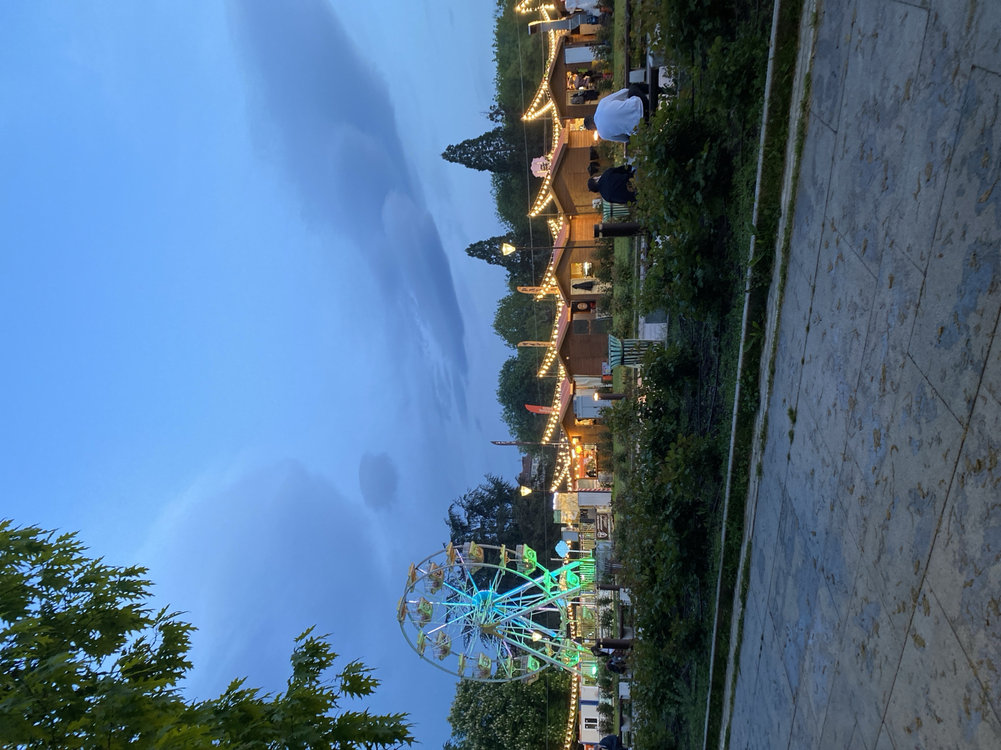 The image captures a Ferris wheel at night, likely located in a park. The sky is a dusky blue with clouds overhead. The Ferris wheel is illuminated in blue, green, and white lights. A row of small wooden buildings or vendor stalls with string lights is visible in the background. A stone-tiled pathway stretches out from the foreground towards the right side of the image. There is greenery on both sides of the pathway, including trees and bushes. People can be seen near the stalls. The specific location in Sofia, Bulgaria, is not determinable from the image.