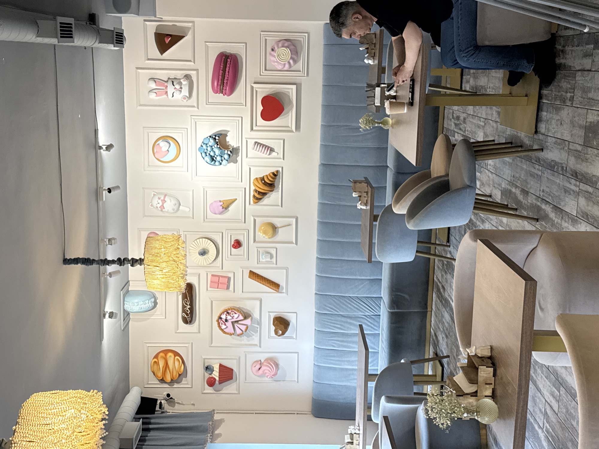 A cafe interior in Saint Petersburg, Russia, features light blue velvet booth seating along one wall, adorned with framed, three-dimensional dessert art. The cafe’s decoration includes chandeliers and framed wall art of assorted desserts. A man stands at a table, partially visible, wearing jeans and a black shirt. Cafe tables are arranged with chairs. Flowers adorn the tables.