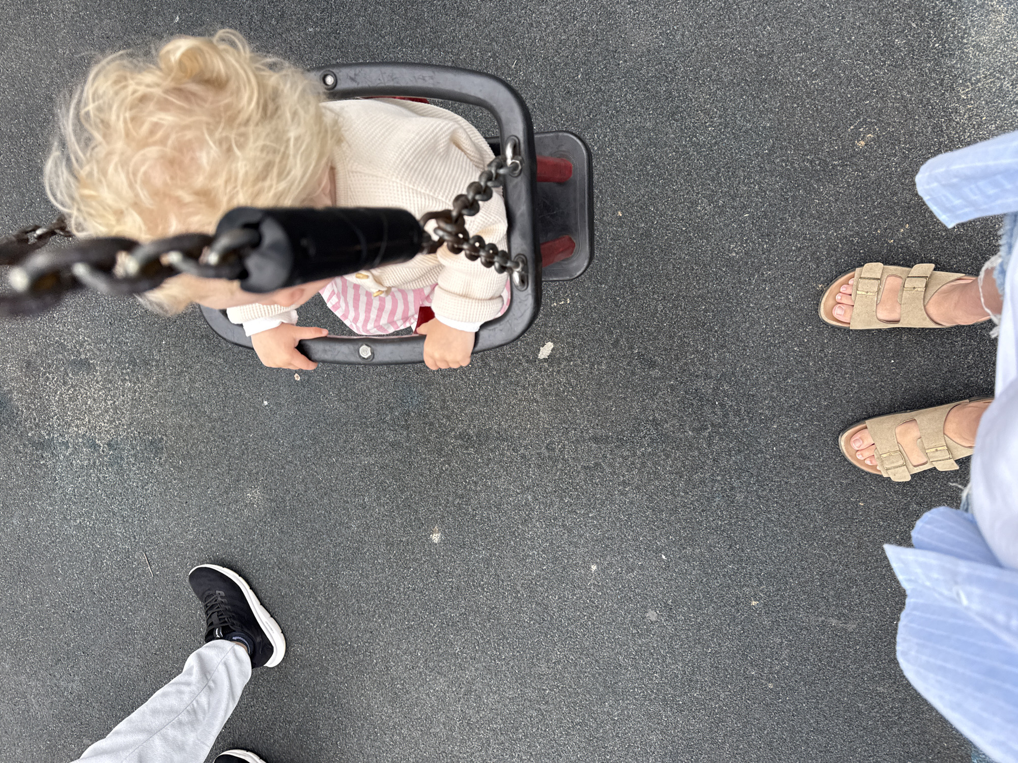 The image is an overhead shot of a child in a swing, with two adults standing on a rubber playground surface. The child, with blonde curly hair, is wearing a cream-colored knitted sweater over a pink and white striped outfit. The child's hands are resting on the black swing's safety bar. One adult is wearing black sneakers and light gray pants, with their feet visible in the lower left corner. The other adult is wearing light brown sandals and a light blue striped shirt, with their feet visible in the upper right quadrant of the image. The scene appears to be a playground, indicated by the rubber surface and swing set. The lighting suggests it is daytime. There is no visible text in the image.