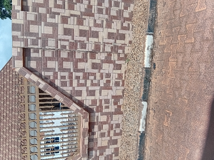 The image displays an exterior scene in Ejule, Nigeria, featuring a residential property boundary. A wall, constructed with brown and beige patterned tiles, dominates the right half of the frame. To the left, a section of a lower wall supports a metal fence, painted a light brown or gold color, featuring decorative scrollwork along the top and vertical bars below. Through the fence, a portion of a building is visible, including a white-framed window and a sloped roof covered in reddish-brown tiles. Above the wall and roofline, a segment of light blue sky with some green foliage is discernible. At the base of the tiled wall, a strip of small, light-colored gravel is present. This gravel strip is bordered by a concrete curb, painted in alternating white and black sections. The foreground consists of reddish-brown interlocking pavers, forming a pathway or road surface. No individuals or active subjects are present in the scene.