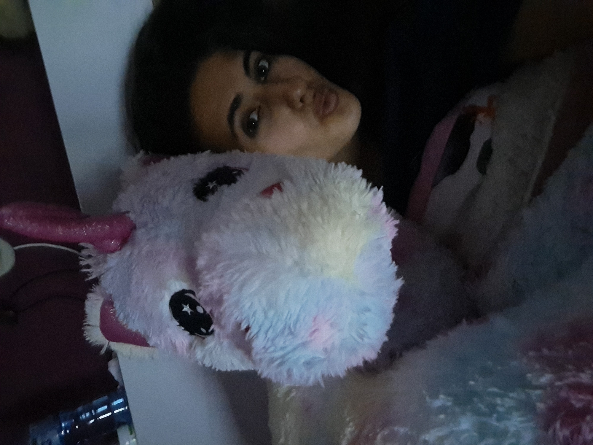 A dimly lit indoor photo shows a young woman or girl lying down next to a large, fluffy unicorn stuffed animal. The image is oriented sideways, with the subjects appearing to lie horizontally across the frame.
The person, positioned in the upper right quadrant of the image, has dark hair and dark eyes. Her face is turned towards the camera, and she is making a "pouty" or "kissing" face with pursed lips. Her expression appears playful or relaxed.
The unicorn plush toy is prominently featured in the lower-left and central parts of the image. It is predominantly pastel-colored, with shades of light pink, light blue, white, and hints of purple, giving it a rainbow-like appearance. The unicorn has large black eyes with white stars printed on them. A pink horn and ear are visible on the left side of the unicorn's head. The texture of the stuffed animal is soft and shaggy.
The lighting is very low, creating a dark background behind the person and making details in the surroundings difficult to discern, though faint outlines of furniture or a wall can be seen. The overall mood is intimate and casual, suggesting a moment of relaxation or play, possibly taken in a bedroom. There is no visible text in the image.
