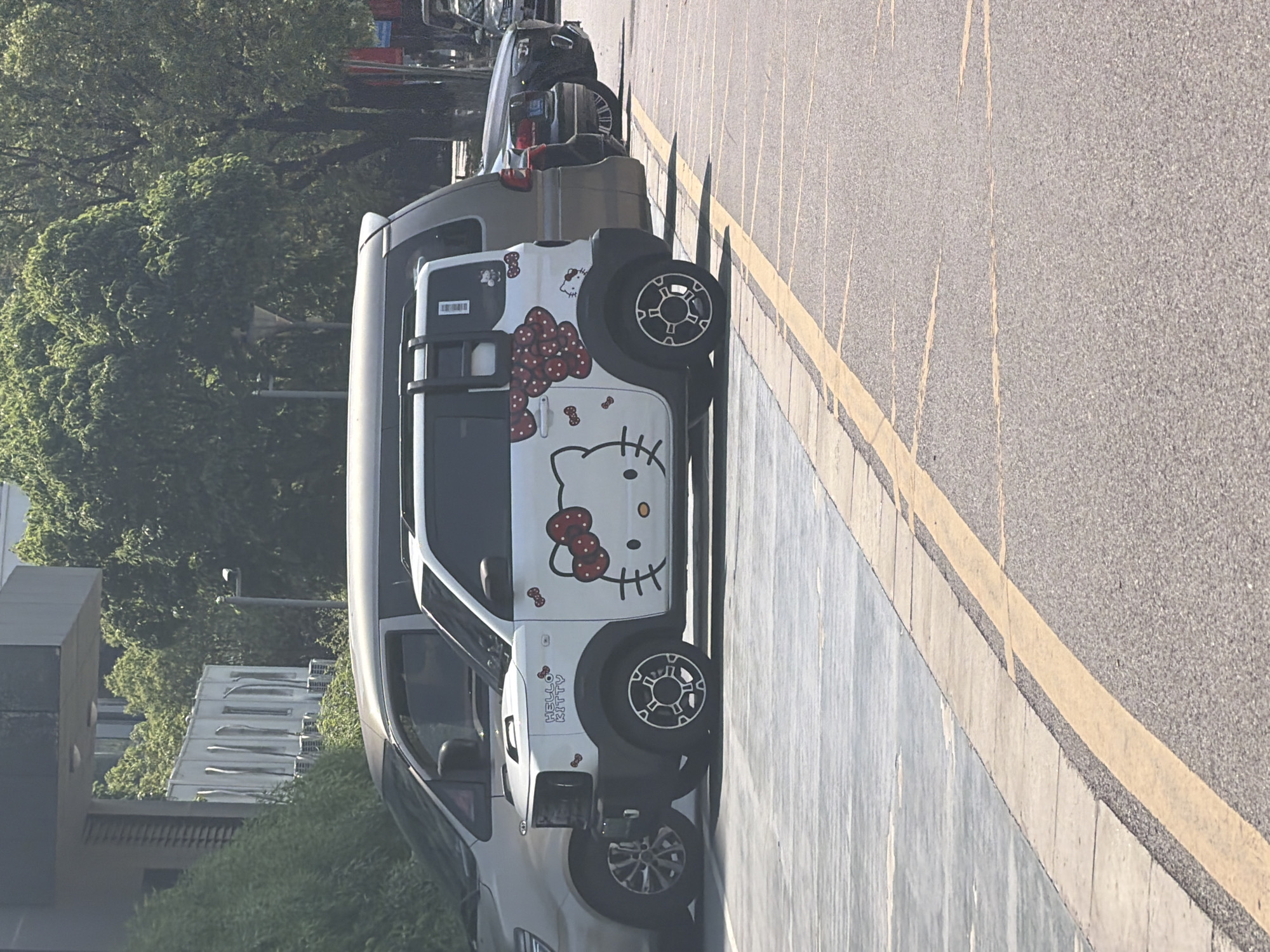 A white miniature electric vehicle adorned with large Hello Kitty graphics, including a prominent face on the side and bows on the rear quarter panel, is parked on an asphalt surface marked with yellow parking lines. The vehicle is positioned in the foreground, partially obscuring a silver minivan to its left and a dark-colored sedan to its right. The environment appears to be an outdoor parking lot, possibly in an urban setting, with a concrete sidewalk and landscaping visible in the background, suggesting it is located in the Jiangning District of China. The lighting indicates a daytime scene, likely under sunny conditions.