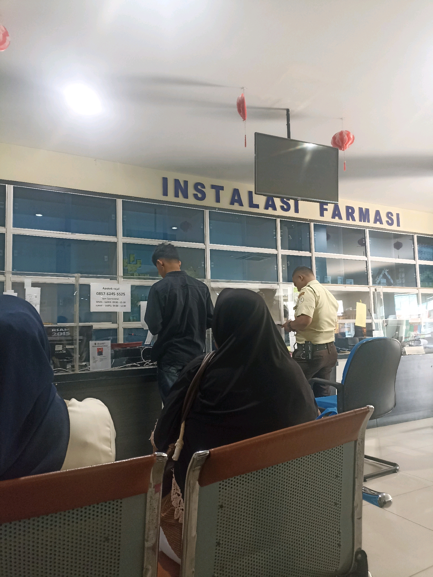 The image depicts the interior of a pharmacy, likely within a hospital or clinic setting, as indicated by the sign "INSTALASI FARMASI" above a counter. The pharmacy is well-lit, with fluorescent lights on the ceiling and a large flat-screen television mounted overhead. Decorative red and white paper lanterns are hanging from the ceiling.
Several individuals are present. In the foreground, three people are seated on chairs facing away from the camera, partially obscuring the view of the counter. One person in the center is wearing a dark hijab. To the left, another person is wearing a dark blue hijab. The third seated person on the right is obscured by the frame.
Behind the counter, two individuals are interacting with staff. A man in a black shirt is standing at the counter, appearing to be engaged in a transaction. Another man, wearing a light brown uniform with a badge, is standing and appears to be a staff member or security guard, interacting with someone at the counter. There are several plastic dividers on the counter.
Various items are visible on the counter and behind it, including what appear to be prescription forms, a computer monitor, a microphone, and shelves stocked with pharmaceutical products. A sign with contact information and operating hours is posted on the counter. The environment suggests a public service area, likely a hospital pharmacy, based on the presence of a uniformed staff member and the overall layout. The specific location in Bandar Lampung, Indonesia, cannot be definitively determined from the visual information alone.