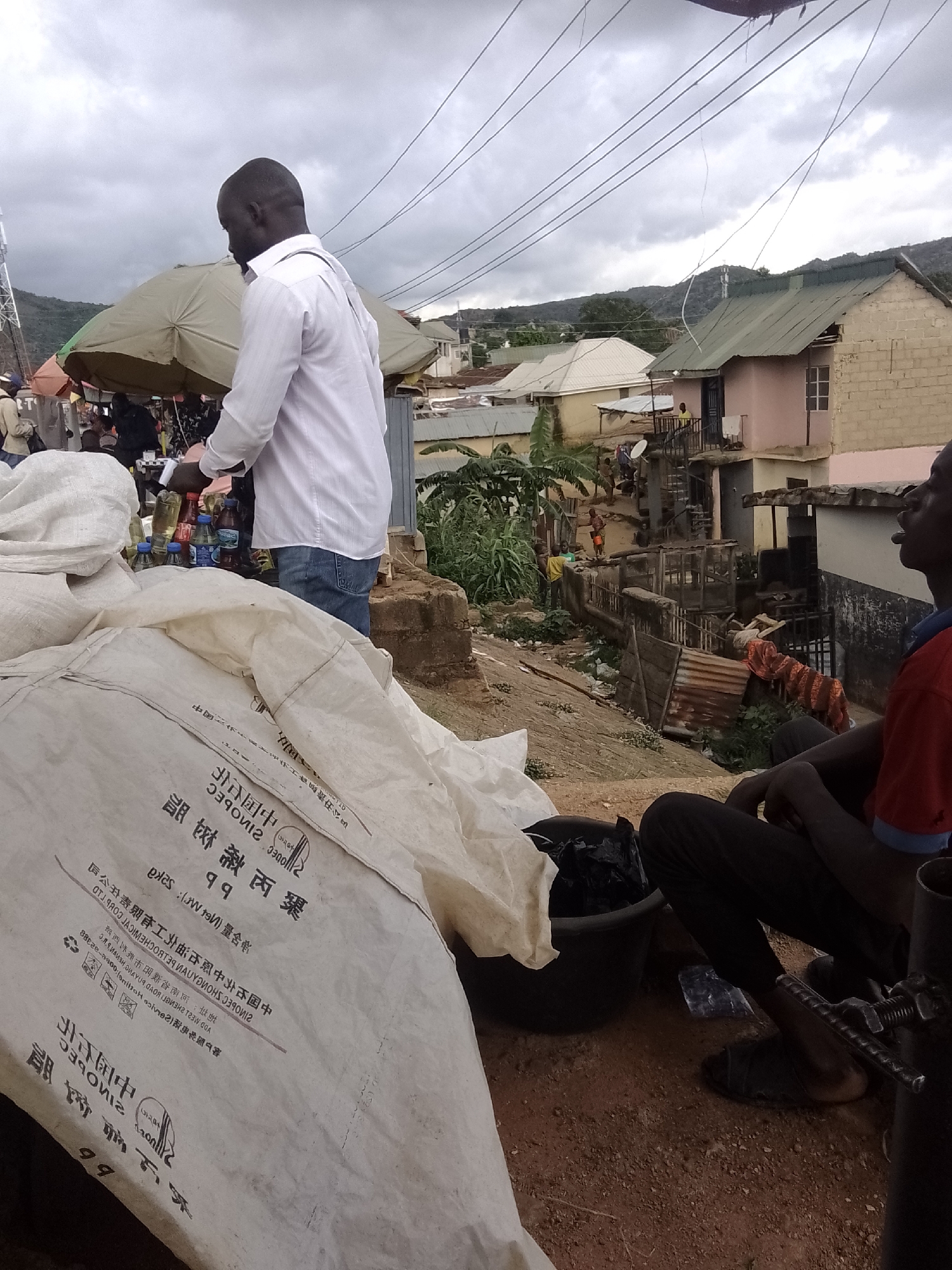This media file captures a street scene with several people and objects. In the foreground, a man in a white shirt and blue jeans is seen from the back, interacting with a stall that has several bottles of beverages. Beside him, another man in a red shirt and black pants is seated, looking towards the right. Large white sacks are piled in the lower left of the frame, with visible text in a foreign language, possibly Chinese, on them.
The background reveals a busy market or residential area with various structures, including houses with tin roofs, some under construction and others appearing older. A large umbrella shades part of the market. People are visible in the middle ground, suggesting a bustling atmosphere. The sky is overcast with clouds, indicating a potentially gloomy or pre-rainy day. The terrain appears to be sloped, with rough ground and some vegetation visible. The overall impression is that of a vibrant, everyday scene in an urban or semi-urban environment, possibly a market. The text on the sacks and potentially on other items in the market hints at international trade or origins.