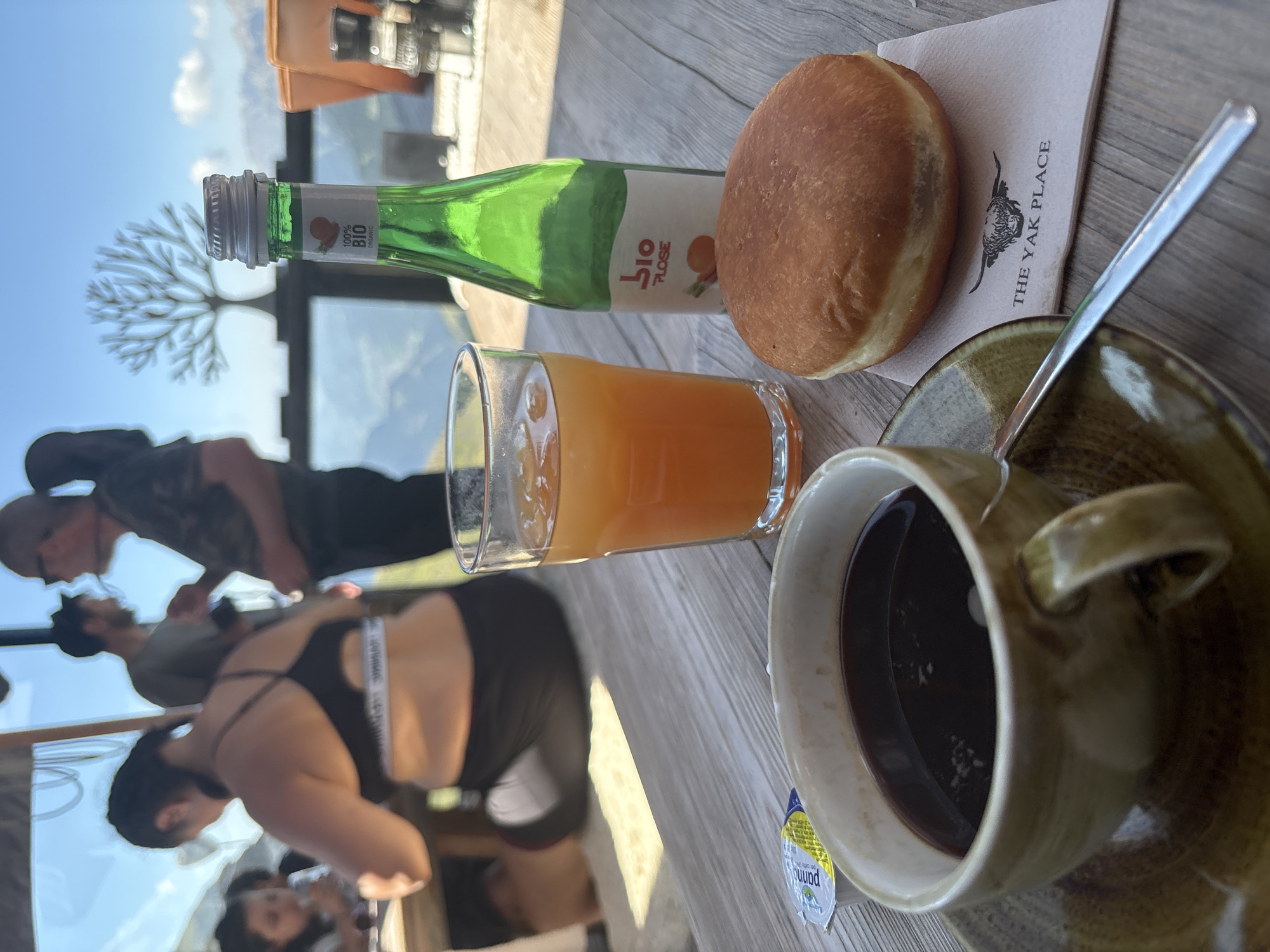 A table setting with food and drinks is pictured outdoors with a scenic mountain view in the background. In the foreground, a brown coffee cup with dark liquid sits on a saucer. To its right, a glass of orange juice with ice is visible. A round pastry with a light brown crust and white filling rests on a napkin with a black yak logo and the text "THE YAK PLACE". A green glass bottle with a white label that reads "100% BIO" is next to the juice. In the background, several people are seated at tables, some blurred. The environment appears to be a restaurant or cafe with a wooden table and chairs, overlooking a mountainous landscape under a clear blue sky. The overall impression is that of a casual dining experience in a mountainous region.