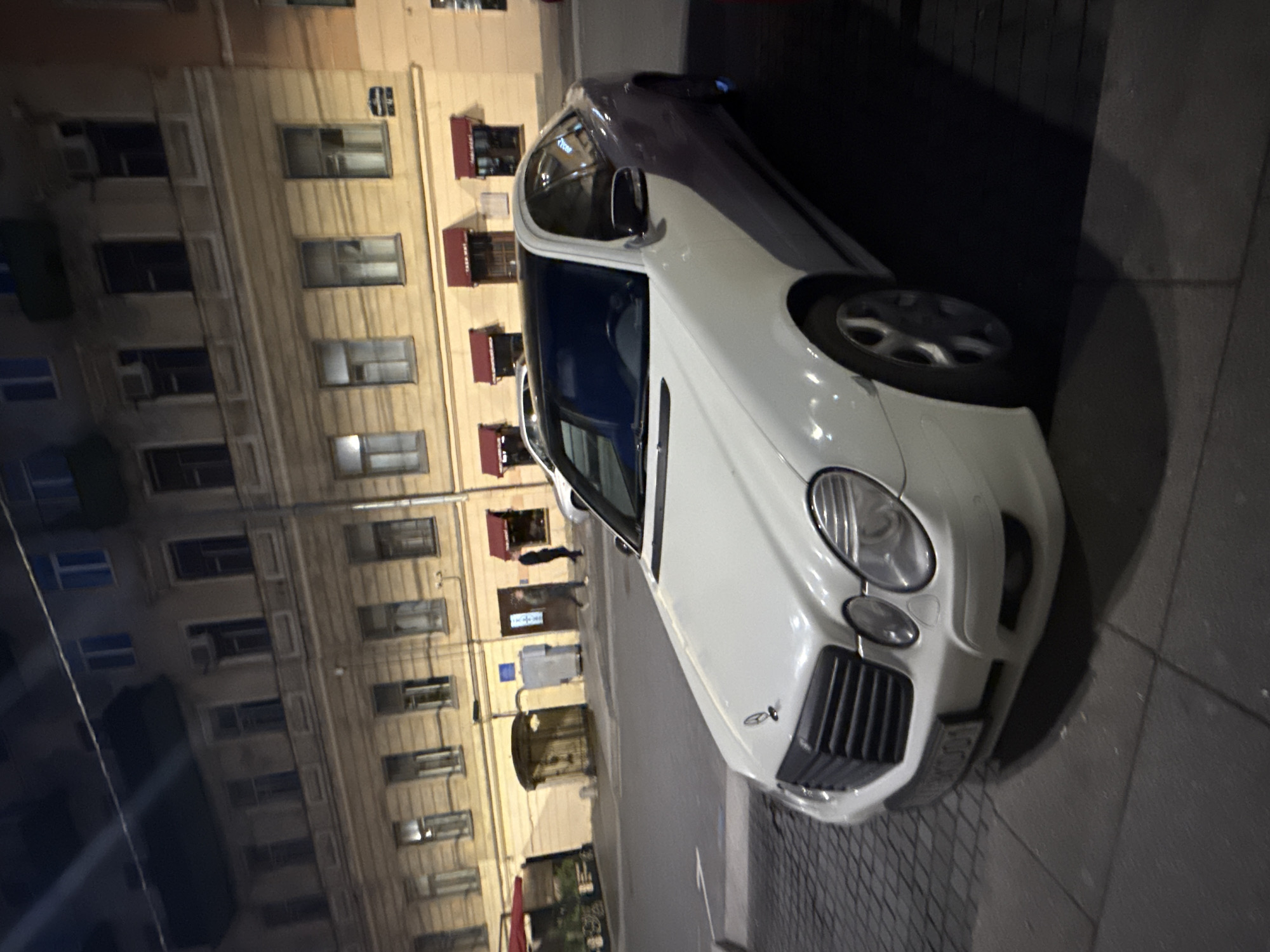 The image captures a white Mercedes-Benz E-Class sedan parked on a cobblestone sidewalk at night on a street in Saint Petersburg, Russia. The car is positioned diagonally, with its front facing the viewer. The background shows a multi-story building with windows and awnings. To the left of the car, there is an entrance to a building, and further to the left, a red awning is visible. Two individuals are seen walking on the sidewalk near the building. One person is walking away from the camera and towards the building entrance, dressed in dark clothing. Another person, also in dark clothing, is walking in the opposite direction. The street appears to be dimly lit, with streetlights casting shadows.