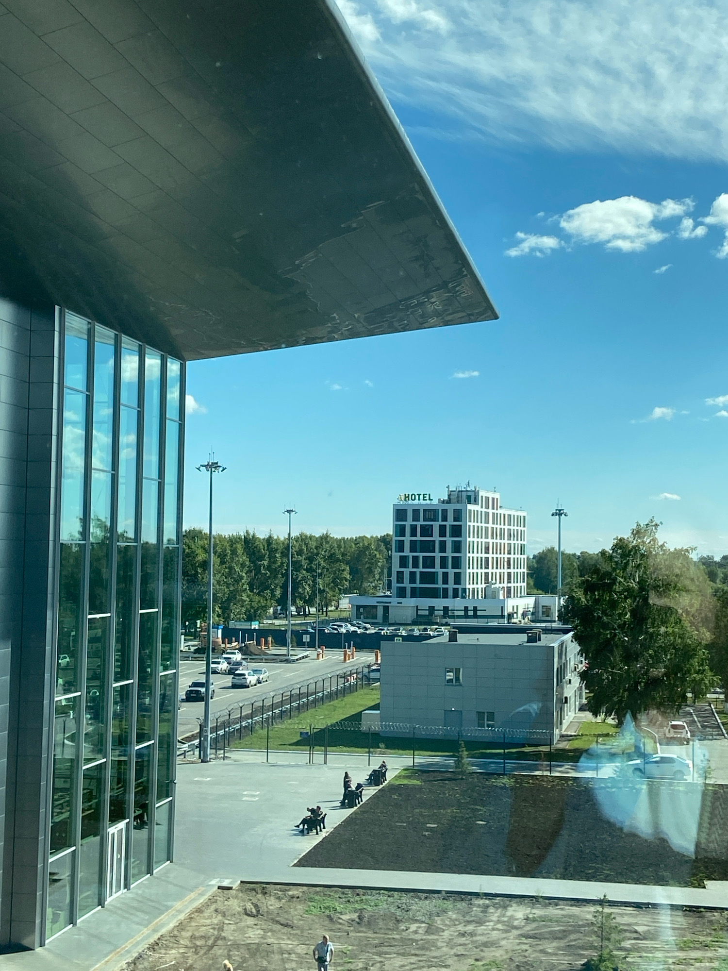 The image depicts an outdoor scene in the city of Ob, Russia. In the foreground, a modern building with a dark facade and large glass windows is visible on the left. A portion of a large, dark, angular overhang from this building extends into the frame from the top left. In the middle ground, there is a paved area with several people seated on benches. Beyond this, a road is visible with cars driving and parked. A fence with barbed wire runs along the side of the road. Further in the background, a multi-story white building with "HOTEL" signage is prominent. Trees and a clear blue sky with scattered clouds complete the environment.