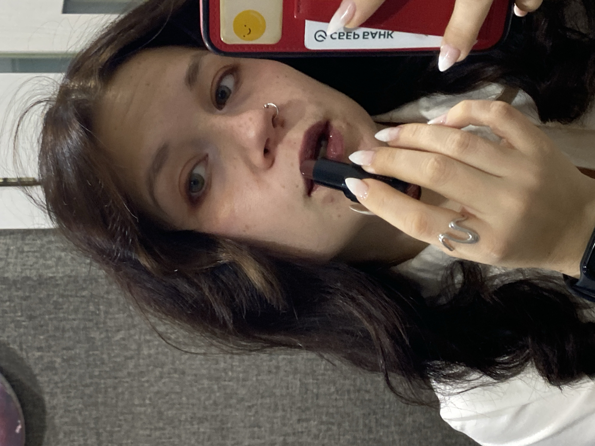 The image shows a close-up of a young woman applying dark lipstick in what appears to be a bathroom or dressing area. She has long, dark, wavy hair, light-colored eyes, and freckles. She is wearing a white collared shirt and a ring shaped like a snake on her right hand. She is holding a black lipstick in her right hand and a red phone case with a cartoon egg sticker in her left hand. The background is a textured gray wall. There is no visible indication of the specific location in Chaikovsky, Russia.