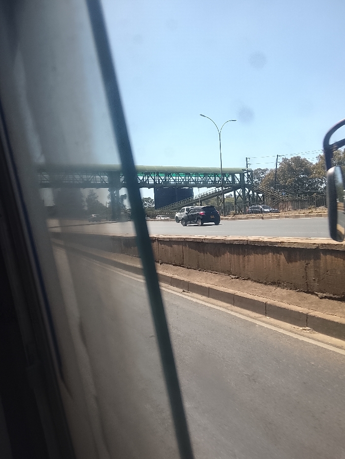 A street scene viewed from inside a vehicle, possibly a bus. A pedestrian overpass spans the road with a tall street lamp nearby. A gray car follows a white van. A low wall separates the road from a bare, sandy area and trees in the background. The sky is a clear, bright blue. Reflections are seen on the interior window of the vehicle.