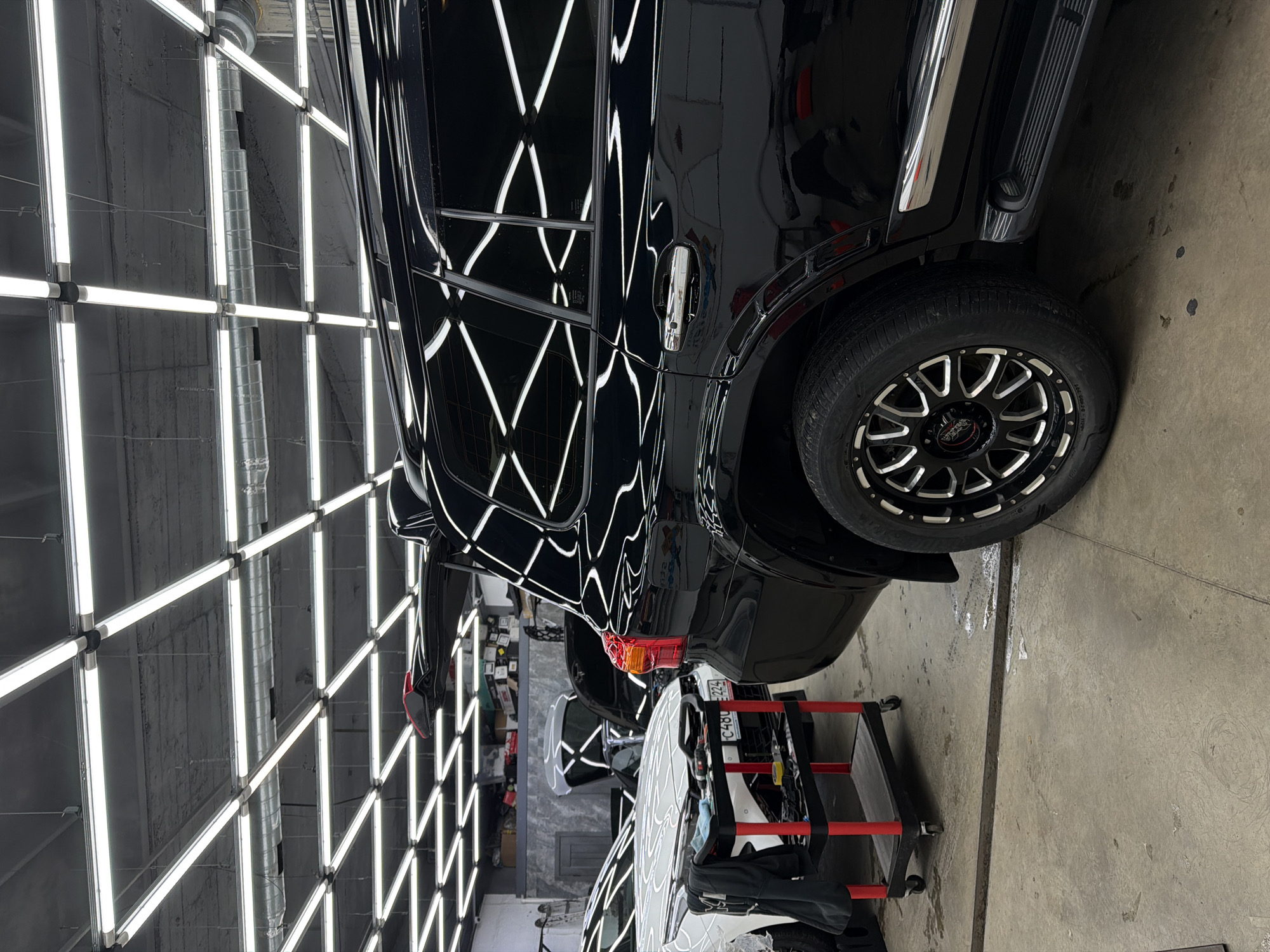 The image depicts a detail shop. A black SUV is being detailed under a ceiling of bright fluorescent lights. The location is in the city of Krasnoyarsk, Russia. A red and black detail cart is next to the vehicle, along with various detailing tools. Another vehicle, possibly a white sedan, is visible in the background.