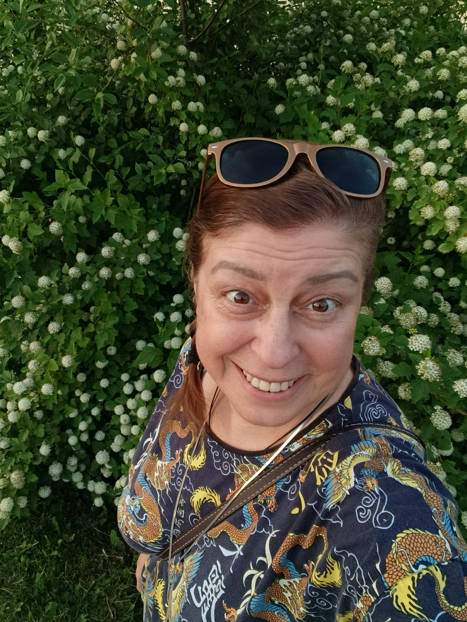 A woman with light brown hair and sunglasses perched on her head is smiling at the camera in front of a dense green bush with small white flowers. She is wearing a dark blue shirt with a dragon and phoenix pattern in orange, yellow, and blue. A brown strap is visible across her shoulder. The background consists of lush green foliage and grass. The scene appears to be outdoors in a park or garden setting. There is no discernible location in the city of Verеya, Russia, as no landmarks or context clues are present in the image to indicate this.