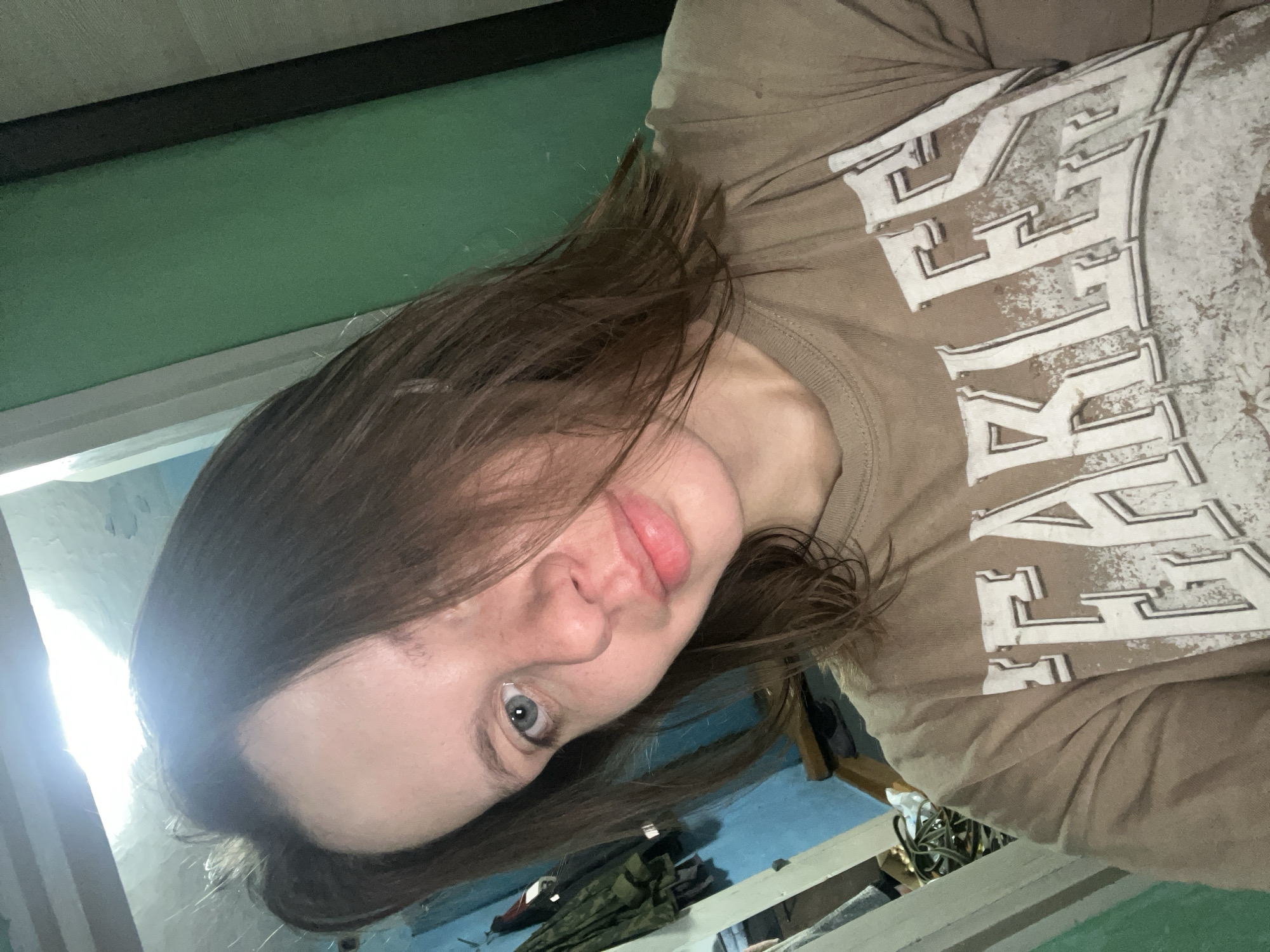 A young woman with medium brown hair and blue eyes is taking a selfie indoors. She is wearing a light brown t-shirt with the word "FEARLESS" printed on it in large white letters. Her hair is partially obscuring her face. The background is cluttered with objects, including what appears to be a blue wall with some hanging items, a green wall, and a wooden structure. There is bright light coming from the top of the image. The location is the city of Nizhny Tagil, Russia.