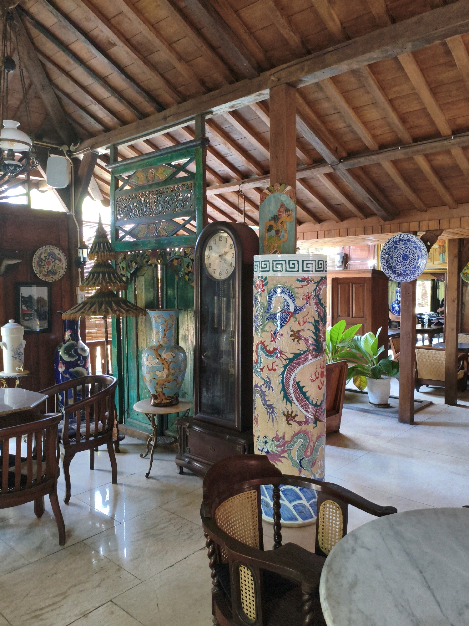 The image shows the interior of a room in Toroh, Indonesia, filled with antique decorative items. The room has a high, wooden, sloped ceiling supported by wooden beams and posts. The walls are also made of wood. In the foreground, several wooden chairs with woven seats and backs surround a marble-topped table. To the left, a tall, white ceramic dispenser with blue floral patterns sits on a small table. Next to it is a dark blue vase with green floral designs. Further back, a large, ornate blue and white vase stands on a small, intricately designed pedestal. To the right of this vase is a tall grandfather clock with a dark wooden casing. Beside the clock, a large, cylindrical ceramic object is decorated with colorful dragon motifs. A potted green plant is visible behind this object. In the background, a dark wooden cabinet and other furniture are visible, along with decorative plates on the wall and a large, intricately carved green wooden screen. The floor is tiled with light-colored marble. The lighting suggests daylight coming from unseen windows.