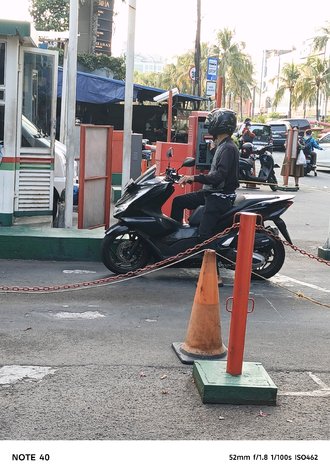 This is a medium shot of a person on a black scooter at a gas station. The person is wearing a helmet and dark clothing. The scooter is parked on the asphalt in front of a red structure with a metal pole. A chain is strung across the foreground. In the background, there is a white building with green trim and other structures. The scene appears to be outdoors, possibly in a city setting. The location is the Pertamina gas station on Jalan Boulevard Raya in Kelapa Gading, North Jakarta, Indonesia.
