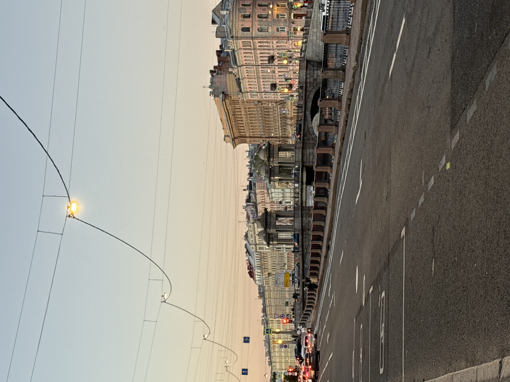 The image depicts a street scene in Saint Petersburg, Russia, likely at dusk or dawn, given the soft, warm light and the sky's gradient from pale blue to peachy orange. A wide asphalt road occupies the foreground, marked with white lane dividers and some parking spaces. Cars are visible in the distance on the road and on a bridge. Pedestrians are present on the sidewalk of the bridge and along the buildings.
The bridge itself is a prominent feature, constructed of stone with ornate balustrades and decorative architectural elements, including what appear to be rotunda-like structures at its ends. Beyond the bridge, a row of historic multi-story buildings lines the street, characterized by their classical architectural style, with intricate detailing, arched windows, and mansard roofs. Some of these buildings display subtle lighting, suggesting they are in use.
Overhead, a network of wires, likely for public transport or utilities, crisscrosses the sky, punctuated by a few hanging streetlights, one of which is illuminated, casting a warm glow. The overall atmosphere is one of urban tranquility, capturing a moment of transition in the day within a historic city. The scene is free of any overt signs of distress or conflict.