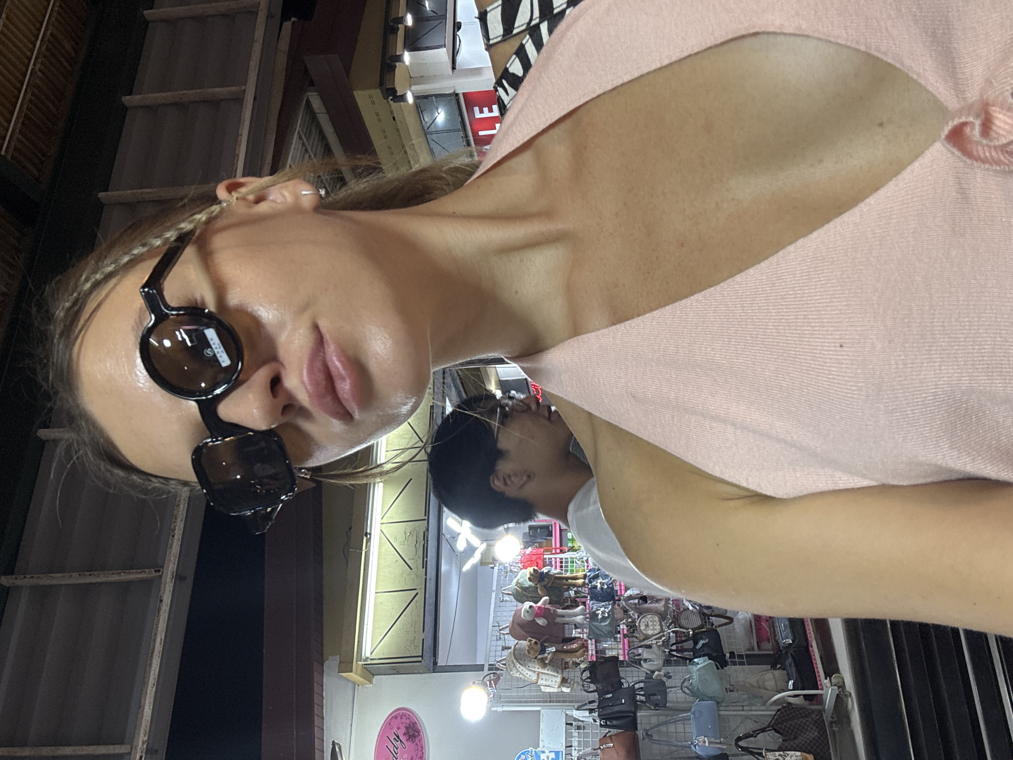 A young woman with blonde braided hair and dark sunglasses is taking a selfie in an outdoor market in Bangkok, Thailand. She is wearing a pink halter top and has a natural makeup look. In the background, a stall is filled with various handbags and backpacks, and another person with dark hair is visible. The scene suggests a casual shopping or tourist experience.