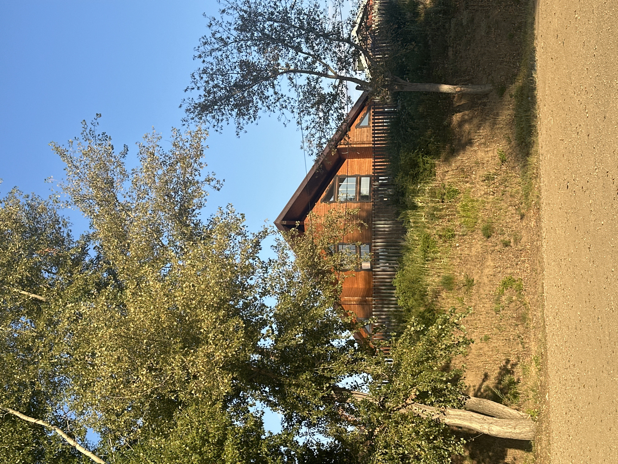 A wooden house with a gabled roof and large windows stands on a grassy embankment behind a wooden fence. Two trees are in the foreground, one with dense green foliage and the other with sparser branches. A dirt road is visible in the bottom of the frame. The sky is clear and blue. The image appears to have been taken in the city of Energetik, Russia.