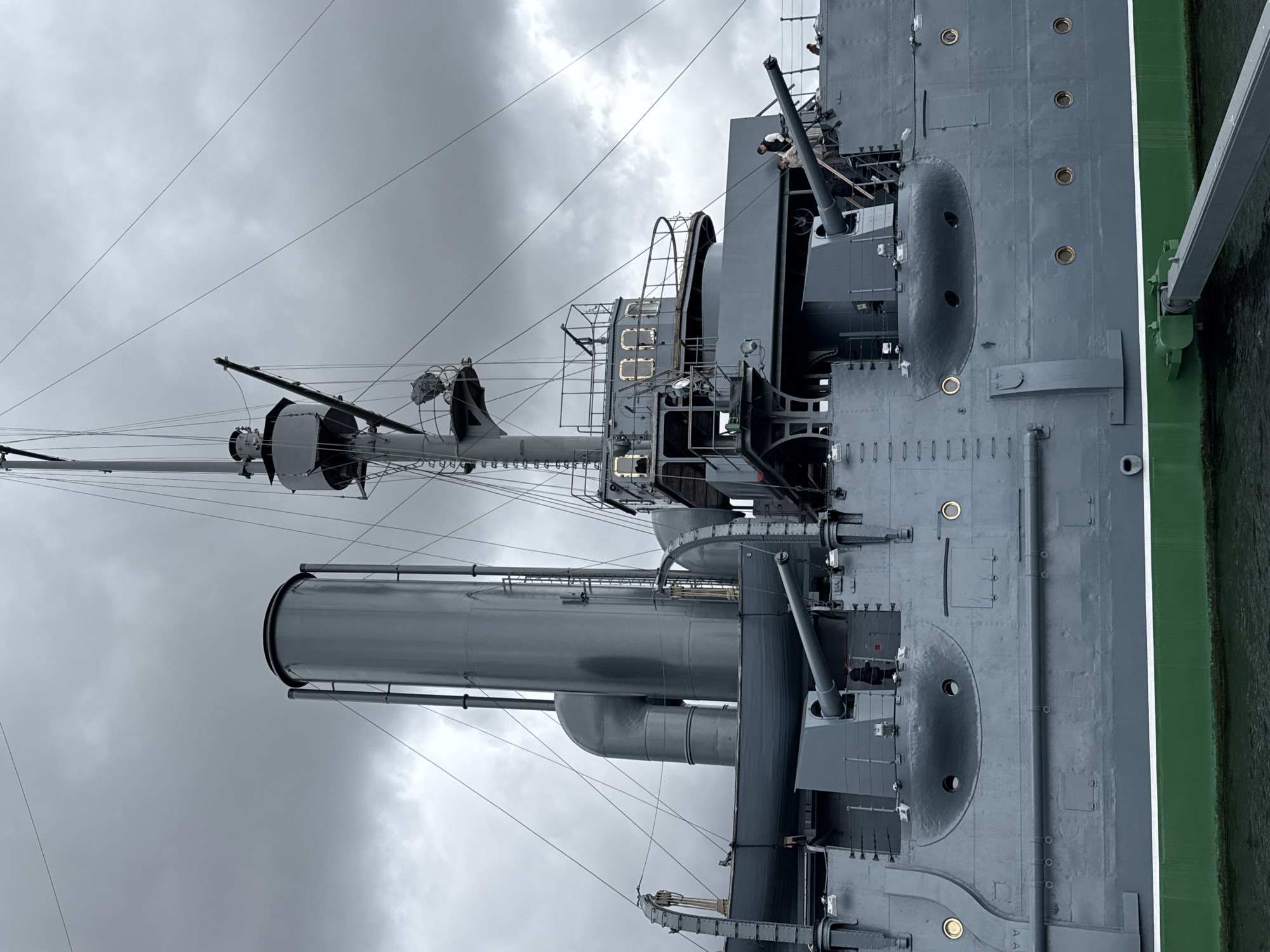 The image depicts the upper structures of the Russian protected cruiser Aurora, a museum ship docked in Saint Petersburg, Russia. The scene captures the ship's masts, smokestacks, gun turrets, and various deck structures against a cloudy sky. Two crew members are visible near one of the gun turrets. The ship is painted in a grey color scheme, and a green strip is seen along the hull. The photograph is taken from a low angle, emphasizing the height of the vessel.