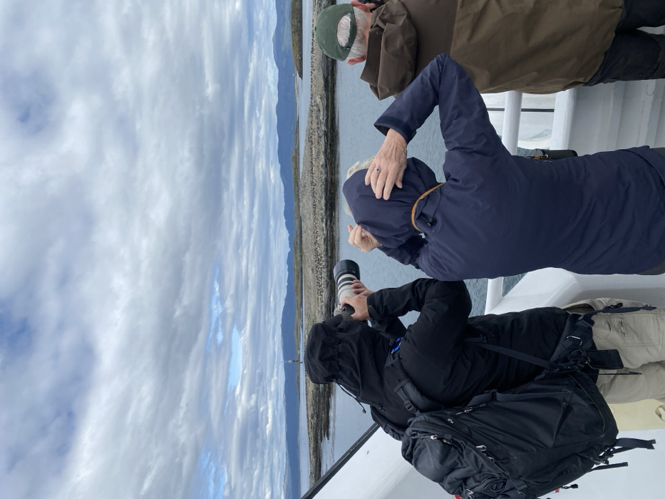 Three people stand on the deck of a boat, gazing at a distant shoreline under a cloudy sky. The foreground shows the individuals are watching something, a person in a black jacket with a backpack takes a photograph with a telephoto lens, another in a blue jacket with their hood up looks toward the shoreline. An older person in a brown jacket and green hat is looking in the same direction.