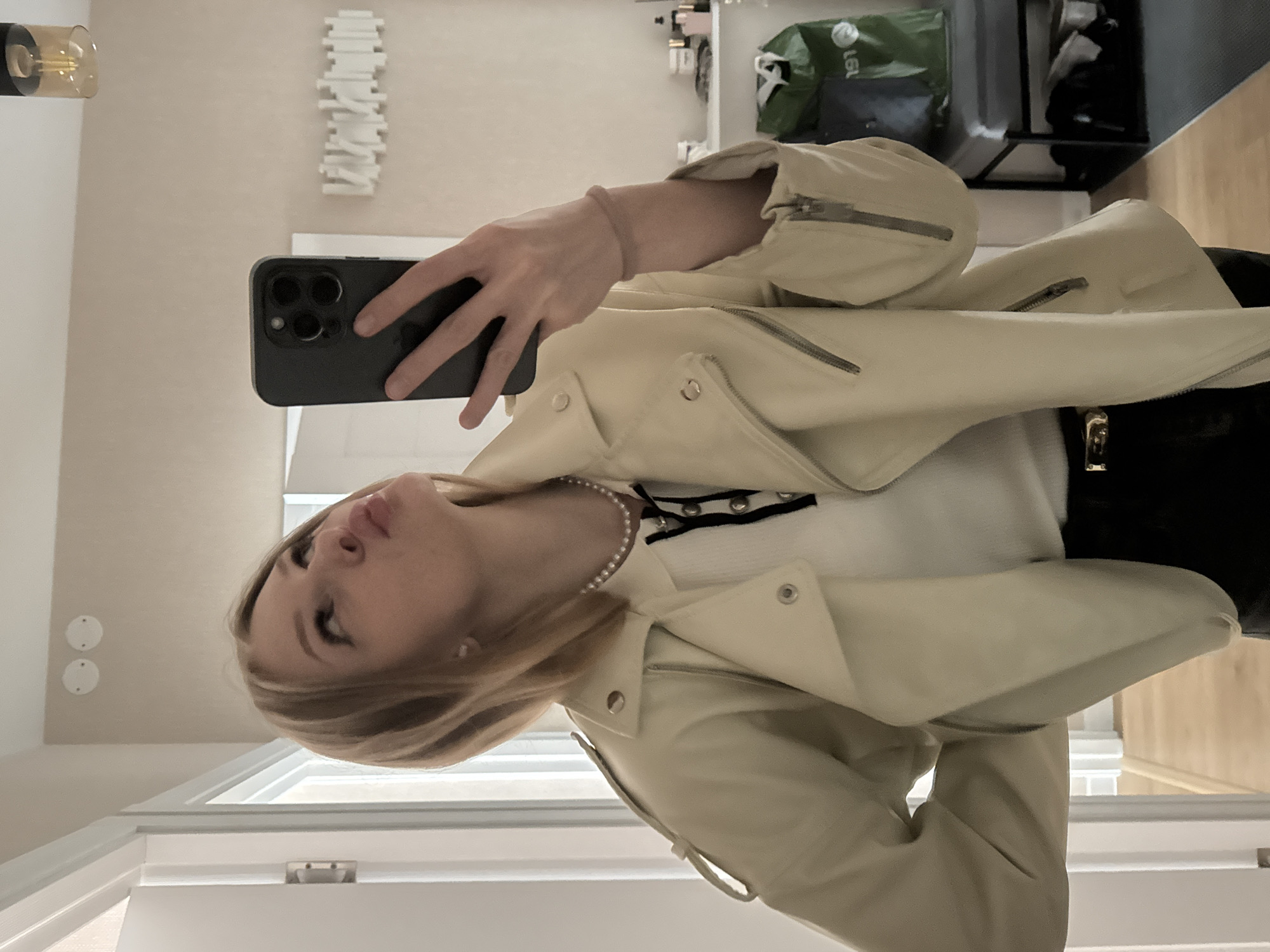 The image depicts a full-length mirror selfie of a young female in an indoor residential setting, typical of a modern apartment or house in the city of Perm, Russia.
The subject, a blonde woman with shoulder-length hair and visible makeup, is positioned primarily on the left side of the frame, holding a dark-colored smartphone with three camera lenses in her right hand to capture the reflection. Her head is tilted slightly back, and her lips are pursed in a stylized expression, with her gaze directed upward towards the ceiling. Her left hand is partially visible on her hip.
She is wearing a cream or light beige faux leather motorcycle-style jacket featuring silver zippers and snaps. Underneath, a white ribbed top with a black lace-up detail at the neckline is visible, complemented by a pearl necklace around her neck. Her lower body is clad in black pants, secured by a belt with a gold buckle. A light pink or beige elastic band is present on her right wrist.
The background, reflected in the mirror, shows a neutral-toned room with light beige walls and white interior doors. Above the subject's head on the left, two circular white ceiling covers are present. To the right, a modern black cylindrical light fixture with a visible amber bulb hangs from the ceiling. Further back on the right, a white shelf holds various bottles and containers. Below this, a dark gray upholstered bench or stool is positioned next to a green reusable bag that partially obscures "191" text. A pile of clothing or items rests beneath the bench. On the wall behind the phone, a white, horizontally oriented decorative piece with a wavy or abstract design is mounted. The flooring transitions from a light wood laminate or parquet in the foreground to a darker gray carpeted area further into the room.