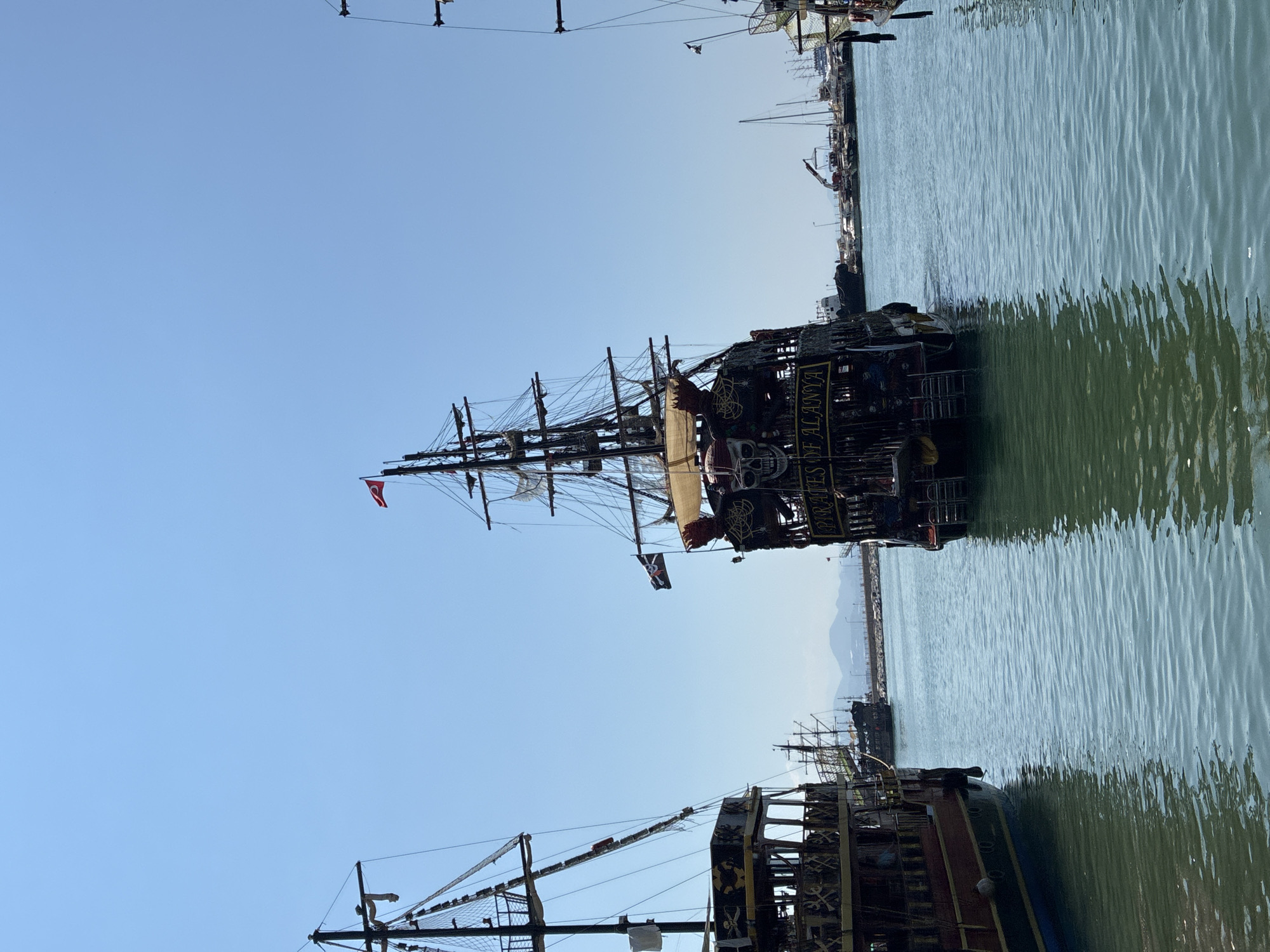The image depicts a harbor scene in Alanya, Turkey, under a clear blue sky. Several large, elaborately decorated pirate-themed ships are prominent in the foreground and middle ground. The ship in the center, with the inscription "PIRATES OF ALANYA," is particularly detailed, featuring skull motifs, rigging, and multiple masts. To the left, another similar pirate ship is visible. In the background, a collection of various other boats, including sailboats and what appear to be smaller tour boats, are docked. Beyond the harbor, a distant coastline with hills or mountains is faintly visible. The water in the harbor is calm and reflects the blue sky and the ships. The overall scene suggests a tourist-oriented harbor, likely a departure point for pirate-themed boat tours in Alanya.