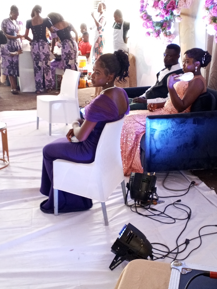 This image captures an indoor or covered outdoor event scene in Ajaokuta, Nigeria. The foreground is dominated by three individuals. A woman on the left is seated in a white chair, wearing a floor-length purple dress with draped shoulders and a pearl necklace, holding a small, light-colored clutch, and looking towards her right. To her right, a man is seated on a dark-colored couch, dressed in a white shirt, black vest, and black bow tie, looking directly forward. Next to him, on the same couch, a woman in a peach-colored, sequined floor-length dress holds a white, possibly illuminated, object near her face and looks towards her right.
The floor is covered with a white fabric sheet. Two black stage lights with visible wiring are positioned on the floor in the mid-ground; one is labeled "FOR LIGHTING."
In the background, multiple individuals are present. Several women are standing, wearing matching patterned purple and black outfits consisting of tops and trousers. Other individuals include a person in a red shirt and another in a white apron. A table with various bottled beverages, including Coca-Cola, is partially visible. The upper right background features floral decorations composed of pink and white artificial flowers arranged on a vertical structure. The overall lighting is bright and even. There are no overt interactions between the main foreground subjects, while background figures appear to be conversing or standing in groups.