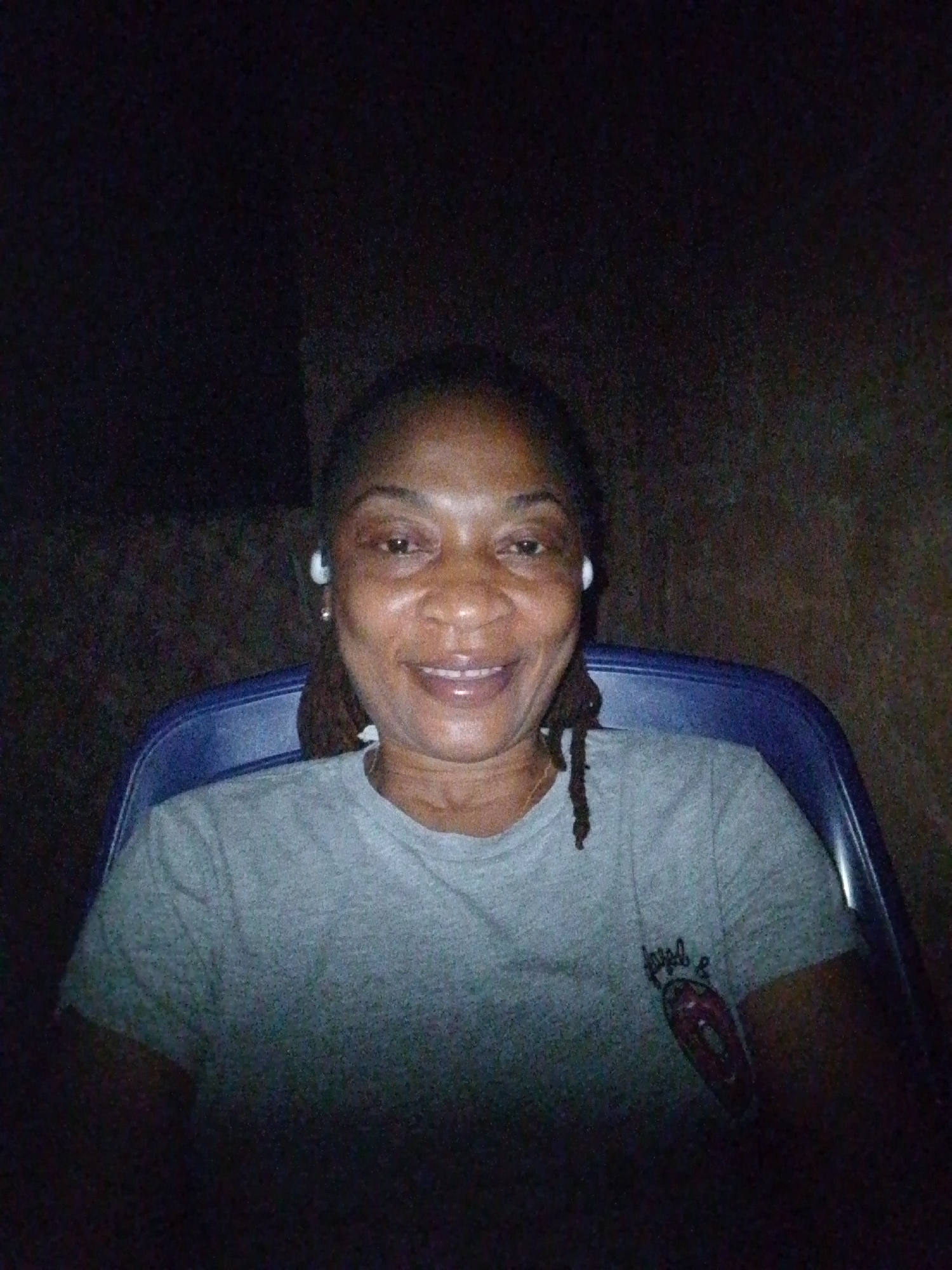 A medium shot captures a woman from the chest up, seated in a blue chair in a dimly lit environment. The woman has dark skin, short dreadlocks pulled back, and is wearing small white earbuds and a grey t-shirt with a graphic on the left chest. She is smiling and looking directly at the camera. The background is dark and appears to be an outdoor setting at night, possibly in Ovoko, Nigeria, given the user's request. The lighting is low, with the main light source appearing to be coming from the front, illuminating the woman's face.