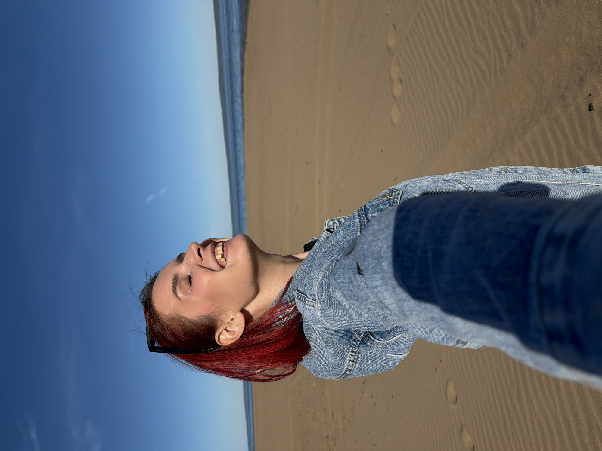 The image depicts a woman with red hair, wearing a denim jacket and sunglasses perched on her head, standing on a beach in Noviy, Russia. Her head is tilted back, and she is laughing. The beach consists of sand, with the ocean and a blue sky visible in the background. The composition suggests she is taking a selfie. Footprints are visible on the sand.