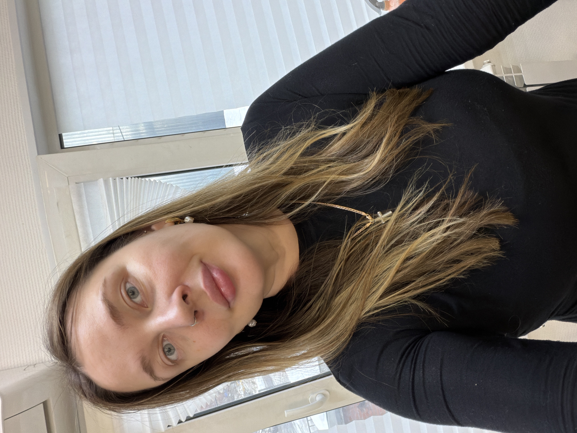 This image depicts a woman captured in a selfie indoors in Chaikovsky, Russia, with the shot taken from approximately the chest up. The subject has long, light brown hair with blonde highlights, parted on the left side, and light-colored eyes. She wears a small silver nose ring in her left nostril, gold hoop earrings with white pearl drops, and a thin gold chain featuring a small gold cross pendant. Her attire consists of a close-fitting black long-sleeved top. She presents a subtle smile, looking directly into the camera.
The background shows a white PVC window frame with white cellular blinds. Through the left pane of the window, a snowy outdoor scene with what appears to be a building facade or railing is visible, indicative of a winter environment. A white window handle is present on the left window frame. To the far right, a partial view of a white horizontal radiator is discernible below the window, and a portion of an orange and black pump-action spray bottle rests on a surface. The interior lighting is even.