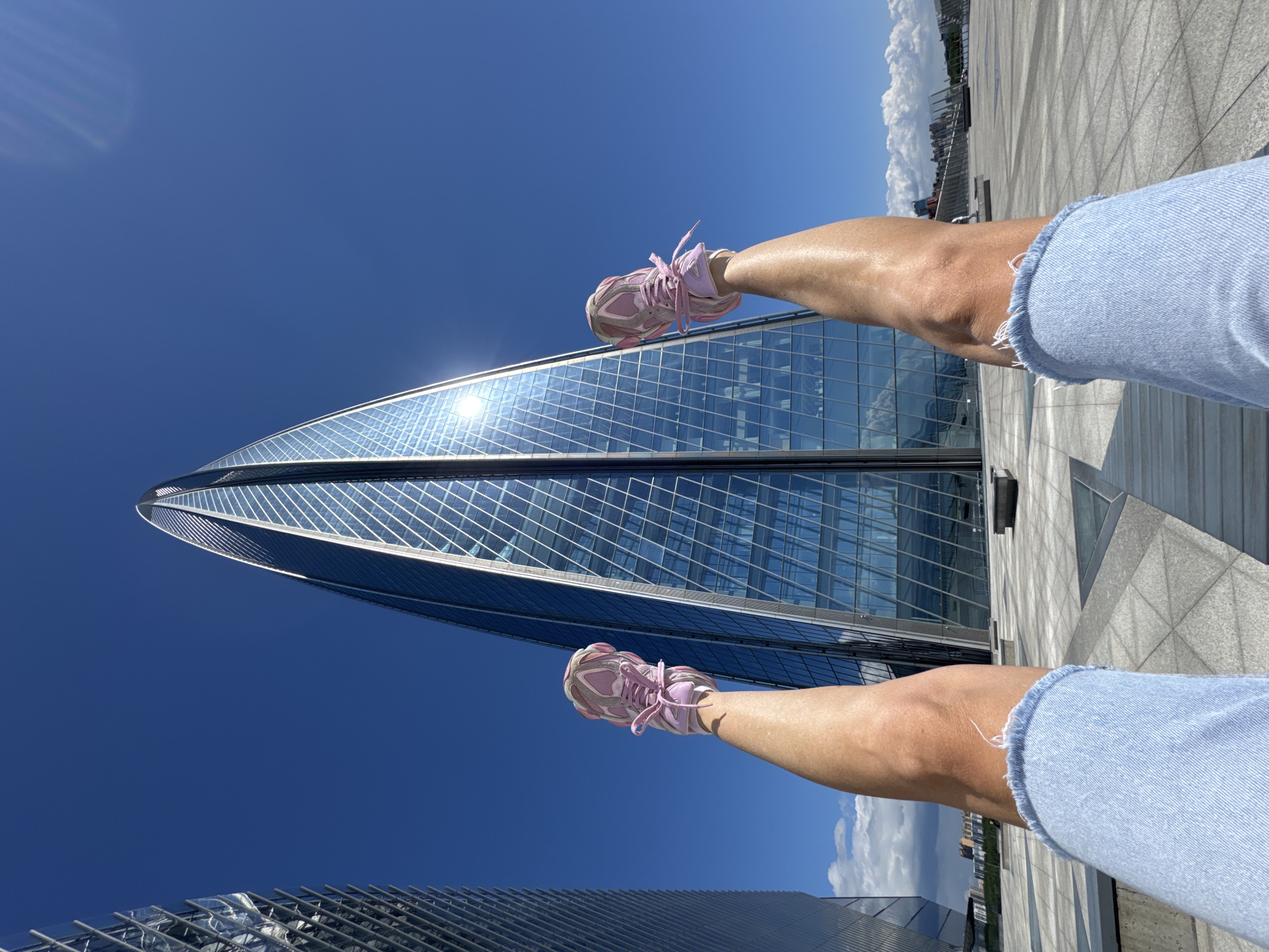 The image displays a person's legs playfully extended against a backdrop of a modern urban landscape under a bright, clear sky. The primary subjects are the lower halves of two legs, from mid-thigh downwards, belonging to a person wearing light blue denim shorts with a frayed hem and distinct pink athletic shoes with white soles and pink laces. The skin on the legs appears tanned.
The scene is set outdoors, likely on a high-level plaza or rooftop in Saint Petersburg, Russia, given the distinctive skyscraper. The most prominent object in the background is the Lakhta Center, a tall, spiraling glass skyscraper with a reflective facade that glints brightly under the sun. The building's sleek, angular lines and glass panels create intricate reflections of the blue sky. The sky itself is a vibrant, cloudless blue, with only a few distant white clouds visible towards the horizon on the right.
The activity depicted is the person lying down or sitting on a paved surface, with their legs raised and extended upwards, creating an engaging, unconventional perspective. The ground below the legs is a light-colored, patterned tiled or concrete surface. The time of day is clearly bright daylight, evidenced by the intense sun reflections and overall illumination. The weather is sunny and clear. No visible text is present in the media file. The composition suggests a relaxed and possibly playful moment captured against an impressive architectural landmark.