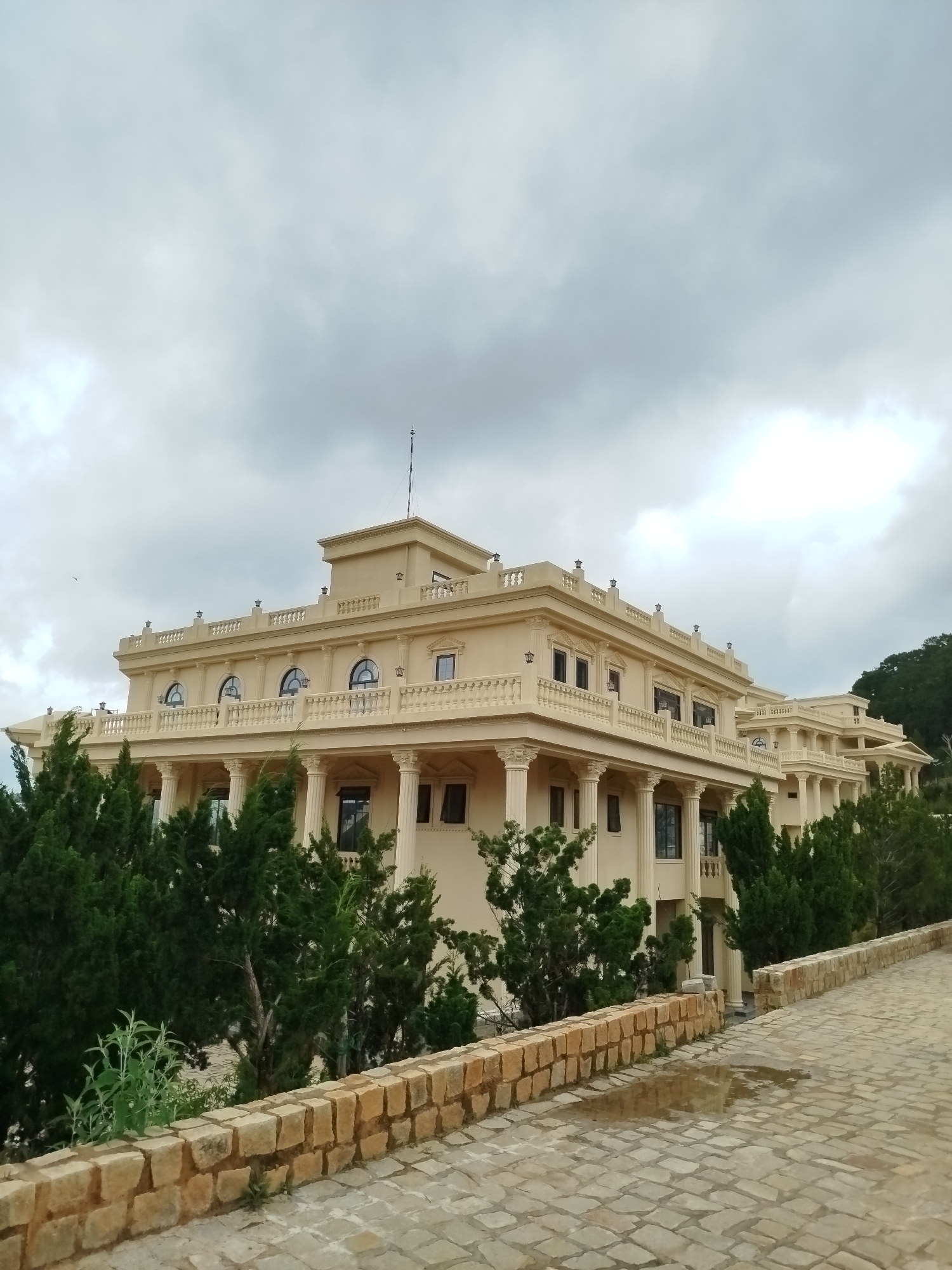 The image shows a grand, cream-colored building with classical architectural elements, including columns, arches, and a balustrade. The building appears to be a multi-story structure with numerous windows and balconies. It is surrounded by lush green trees, some of which are tall and slender, while others are shorter and bushier. In the foreground, there is a cobblestone path that leads up to the building. The path is bordered by a low stone wall made of rough-hewn blocks. A small puddle of water is visible on the path in the lower right portion of the image. The sky above is overcast with heavy clouds. The overall impression is one of opulence and grandeur set against a natural, slightly moody backdrop. While the location is stated as Lạc Dương, Vietnam, there are no specific visual cues in the image that definitively confirm this geographical location, such as signage or distinct local architecture beyond the building itself.