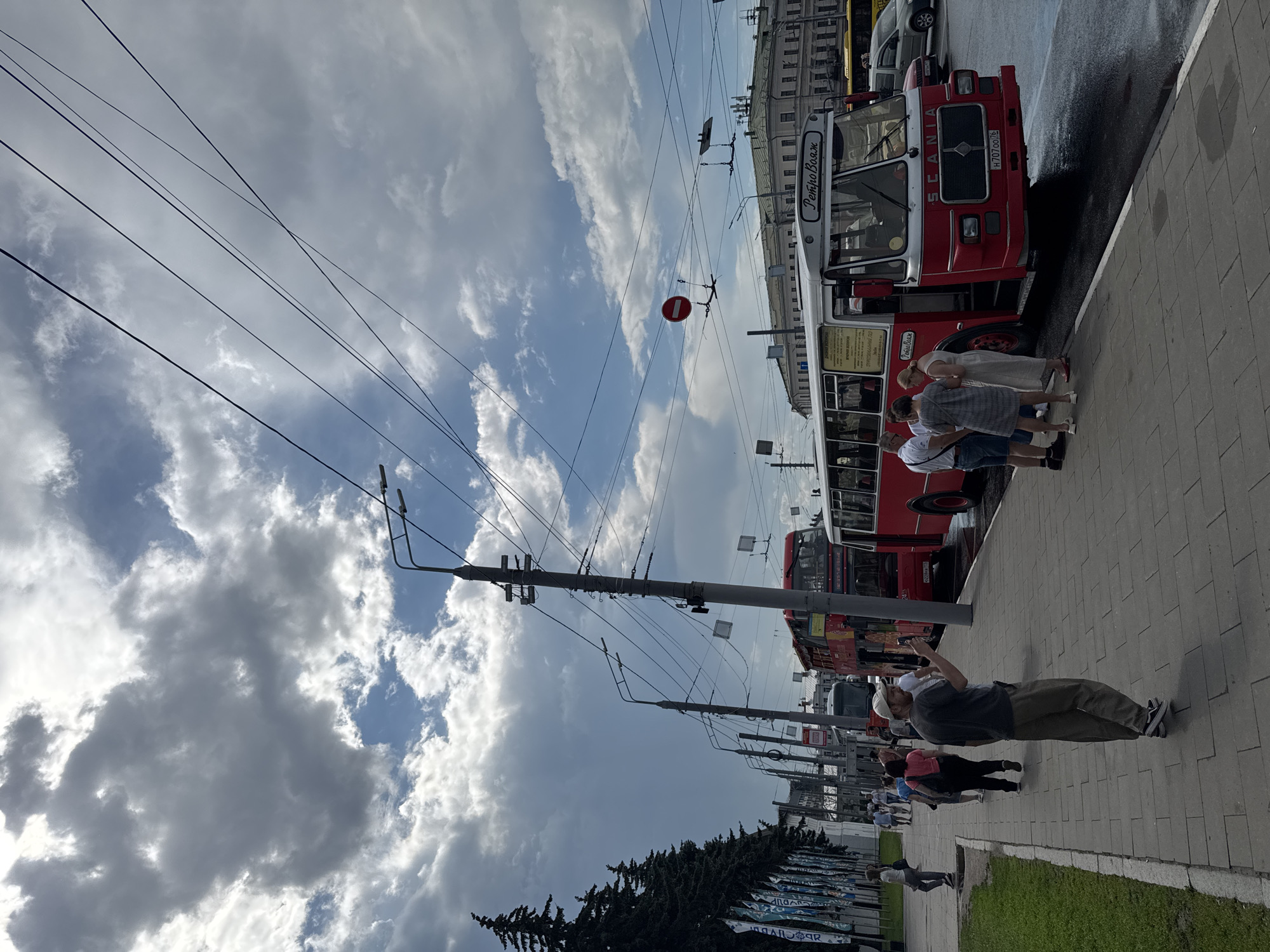 A group of people are standing on a sidewalk next to a red and white bus in Заволжье, Russia. The bus has the word "РетроВояж" written on the front. The sky is cloudy.