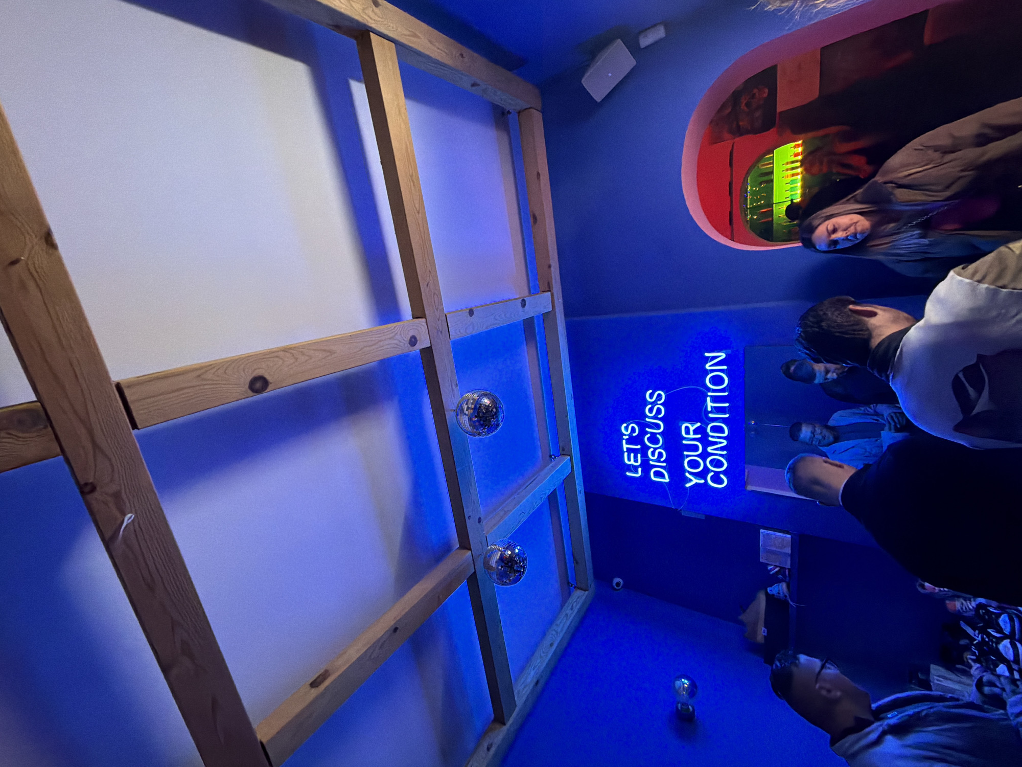 The image captures an indoor scene featuring a blue-lit space with a wooden framework in the foreground, decorated with disco balls. In the background, there's a group of people, some partially visible, appearing to be engaged in conversation. A red-rimmed circular opening can be seen above them, offering a glimpse into another area. The text "LET'S DISCUSS YOUR CONDITION" is displayed in blue neon lighting on the wall. The setting is likely a bar or an entertainment venue, potentially in Sofia, Bulgaria, but specific landmarks are absent, making precise location confirmation difficult.