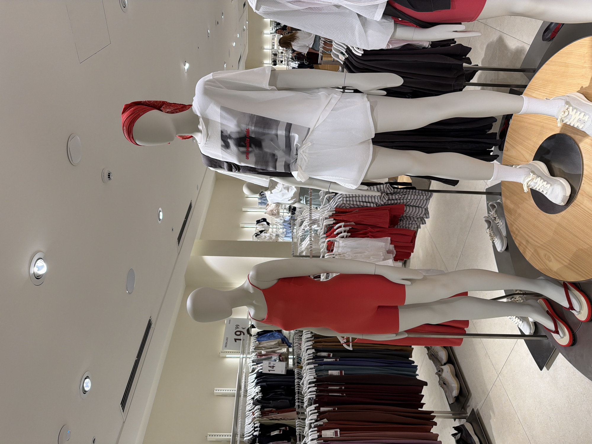 The image shows a retail store interior, likely a clothing store. In the foreground, two mannequins are displayed. The mannequin on the left is wearing a bright red, sleeveless, one-piece garment that appears to be a leotard or romper, along with red and white flip-flops. The mannequin on the right is wearing a white t-shirt with a black and white graphic image on the front, white shorts, and white socks with white sneakers. This mannequin also has a red bandana tied around its head. Both mannequins are standing in front of racks of clothing. The store environment features bright overhead lighting, white ceilings, and light-colored walls. Racks of clothing are visible in the background, displaying a variety of garments in different colors and styles. Some price tags are visible on the clothing racks, with one indicating "19'99 €" and another "14'99". The overall impression is a well-lit, organized retail space. The image is consistent with a store located in a shopping center, such as the La Marina shopping center in Finestrat, Spain, given the style of the store and the presence of Spanish currency.