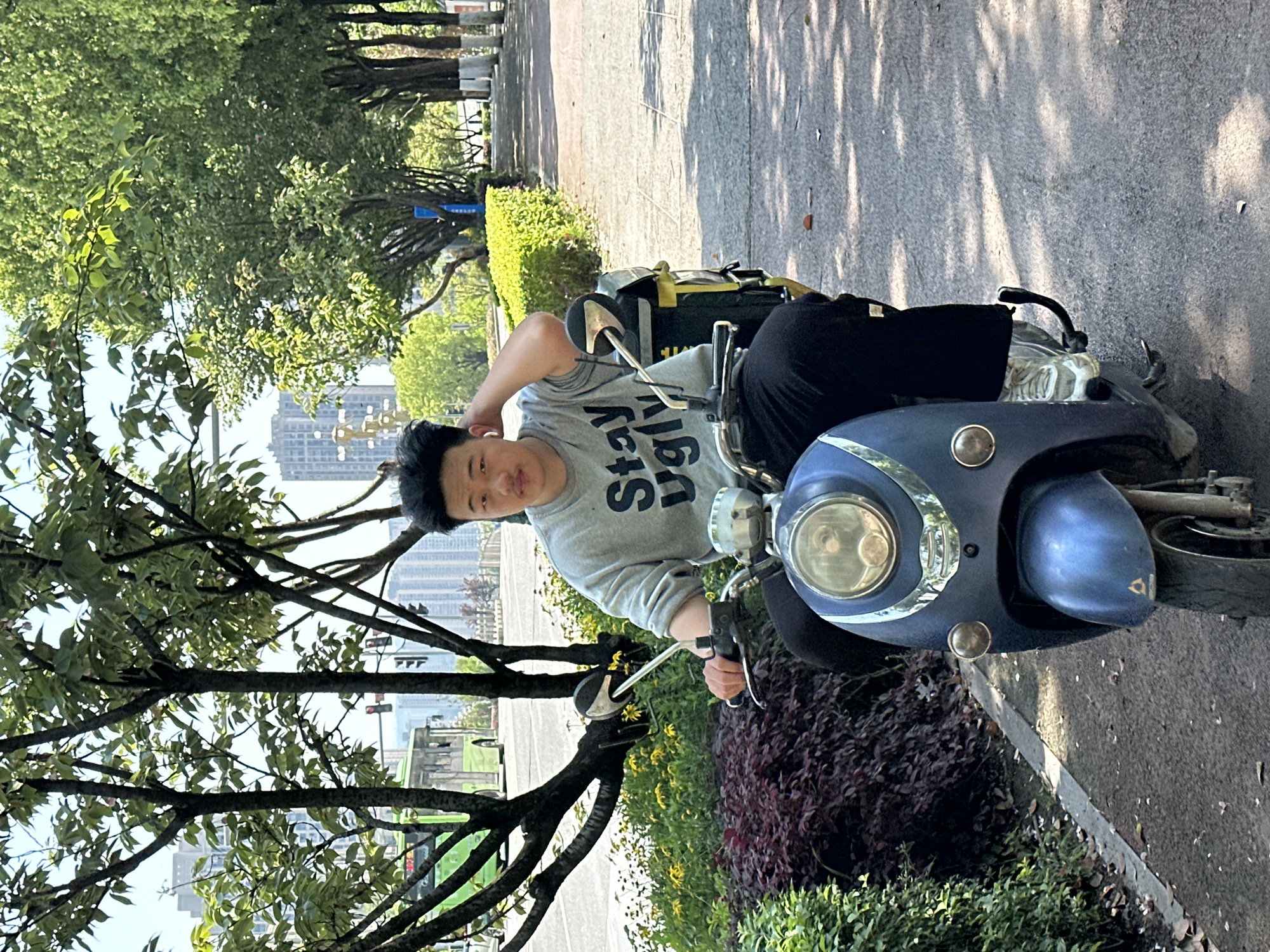 A man is sitting on a blue moped on a paved path next to a green hedge and a dark purple shrub. The man is wearing a gray sweatshirt with the words "Stay Ugly" and black pants. He has his right hand on his head and is looking at the camera. Behind the hedge is a row of trees, and behind the trees are buildings. A green bus is visible between the trees. The location is Jiangjin, China.