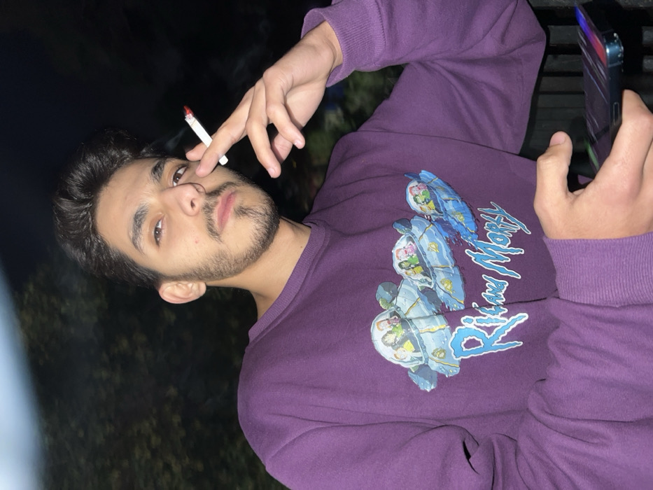 The image features a young man, likely in his late teens or early twenties, photographed at night in Al-Muhajerin Municipality, Syria. He has dark hair, a neatly trimmed beard, and a mustache, and is looking directly at the camera with a composed, neutral expression.
He is wearing a long-sleeved purple sweatshirt adorned with a prominent graphic design. The design includes the stylized text "Rick and Morty" in a light blue font, accompanied by several cartoon characters from the series depicted within futuristic, helmet-like capsules or small spacecraft, rendered in various shades of blue, green, and white.
The man's right hand holds a lit cigarette between his index and middle fingers, with the glowing red tip clearly visible. His left hand, visible on the right side of the frame, is holding a smartphone, which appears to be an iPhone given its characteristic design elements. The phone's screen is partially visible and illuminated.
The background is very dark and out of focus, suggesting the photo was taken at night outdoors, possibly with a flash, as the subject is well-lit. The overall scene conveys a casual setting, with the man engaged in smoking and holding his phone.
