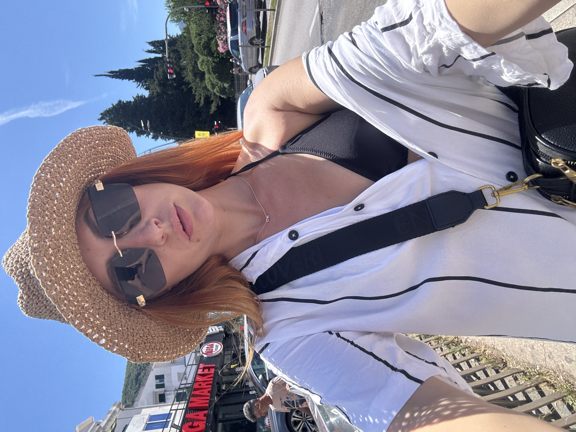 A woman with auburn hair, wearing a straw hat, oversized sunglasses, a black bikini top, and an open white and black striped shirt, stands on a sidewalk in Budva, Montenegro. She is holding a black crossbody bag with gold hardware. Behind her, a street is visible with traffic, trees, and buildings, including a sign for "MEGA MARKET." The sky is clear blue with a faint cloud trail. The woman is taking a selfie.