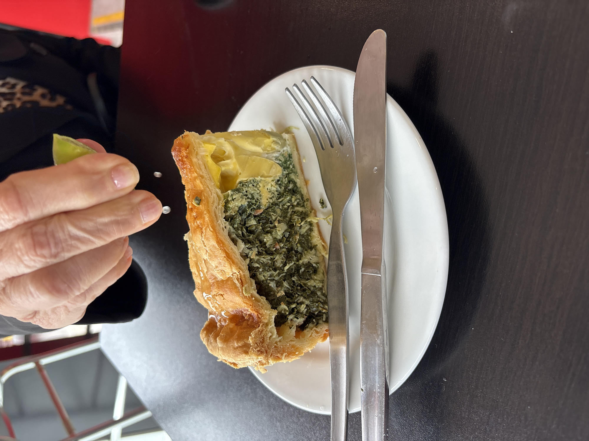 A high-angle shot shows a plate of spinach quiche with a fork and knife. A person’s hand is squeezing a lime wedge. The scene is set on a dark table with hints of a red railing and red-and-white chairs in the background, suggesting an outdoor or semi-outdoor eating area. The location is most likely San Luis, Peru.
