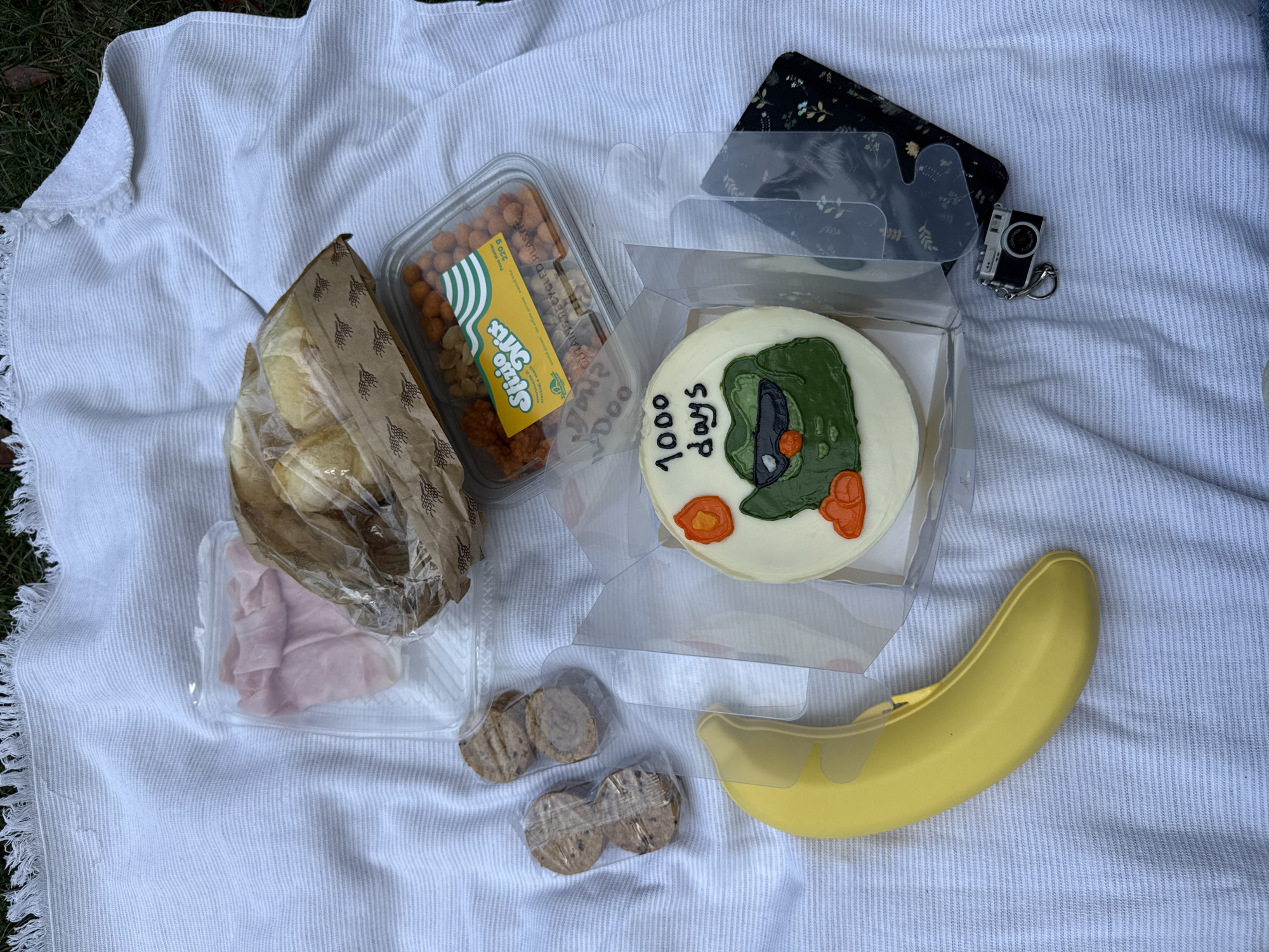 The image shows a picnic spread laid out on a white striped blanket on grassy ground. The spread includes a plastic container of sliced ham, a brown paper bag containing what appear to be bread rolls, a clear plastic container filled with mixed nuts, a yellow banana, a small round cake decorated with a frog wearing sunglasses and the text "1000 days" written in black icing, and a small keychain shaped like a vintage camera. There are also two small plastic-wrapped packages containing round cookies or biscuits. The background suggests an outdoor setting. No specific location in Milan, Italy is identifiable from the image.