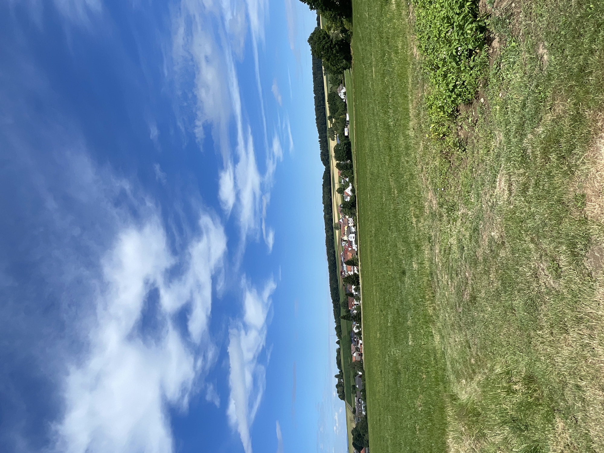 An outdoor, eye-level, vertical, medium shot captures a sunlit rural landscape under a bright blue sky with scattered white clouds. In the foreground, a vast, green field with patches of dry, brown grass dominates the scene. A faint path is visible on the left side of the field. In the middle ground, a village with red-roofed houses and white facades is nestled amongst trees and rolling hills. The village appears to be Christerode, Germany. The background features a dense line of dark green trees atop the hills, with a lighter green and yellow field visible behind the village. The overall impression is one of a serene, pastoral environment.