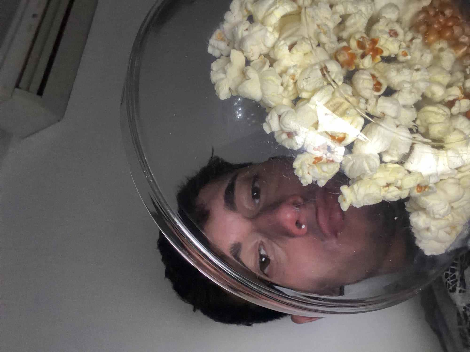 A low-angle shot shows a clear bowl filled with popcorn. Reflected in the bowl is a person’s face. The face is male with dark hair and a piercing on the left nostril. The background is a plain, light-colored wall. There is no information to indicate this photograph was taken in Blagoevgrad, Bulgaria.
