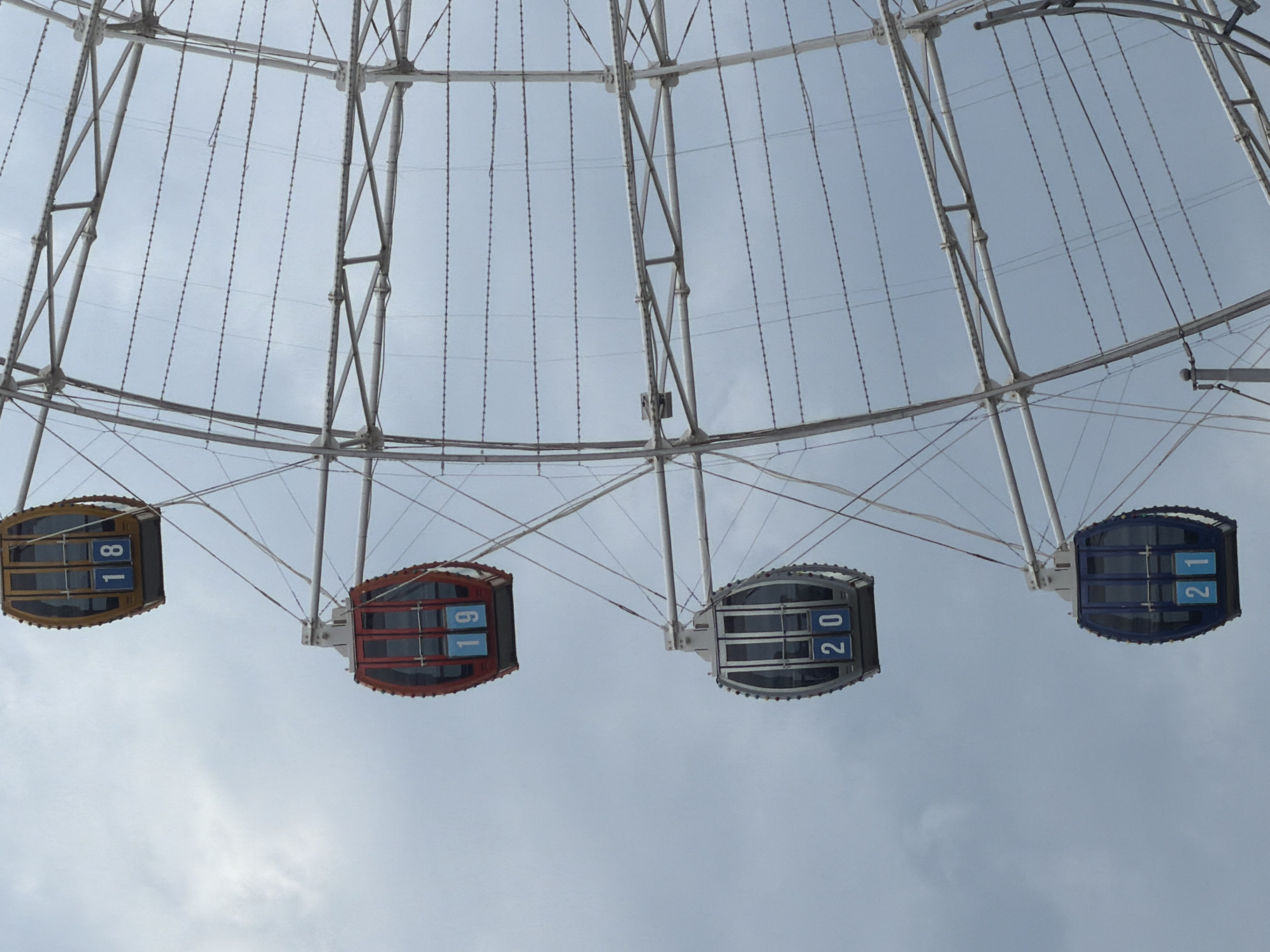 The image shows a close-up view of a Ferris wheel against a cloudy sky. Four gondolas are visible, numbered 18, 19, 20, and 21. The gondolas are enclosed with windows and have blue panels displaying the numbers. The Ferris wheel structure is made of white metal beams and cables. There are no people visible in the image. The scene appears to be from an amusement park or fairground. This Ferris wheel is located in Astana, Kazakhstan, specifically in the Presidential Park.