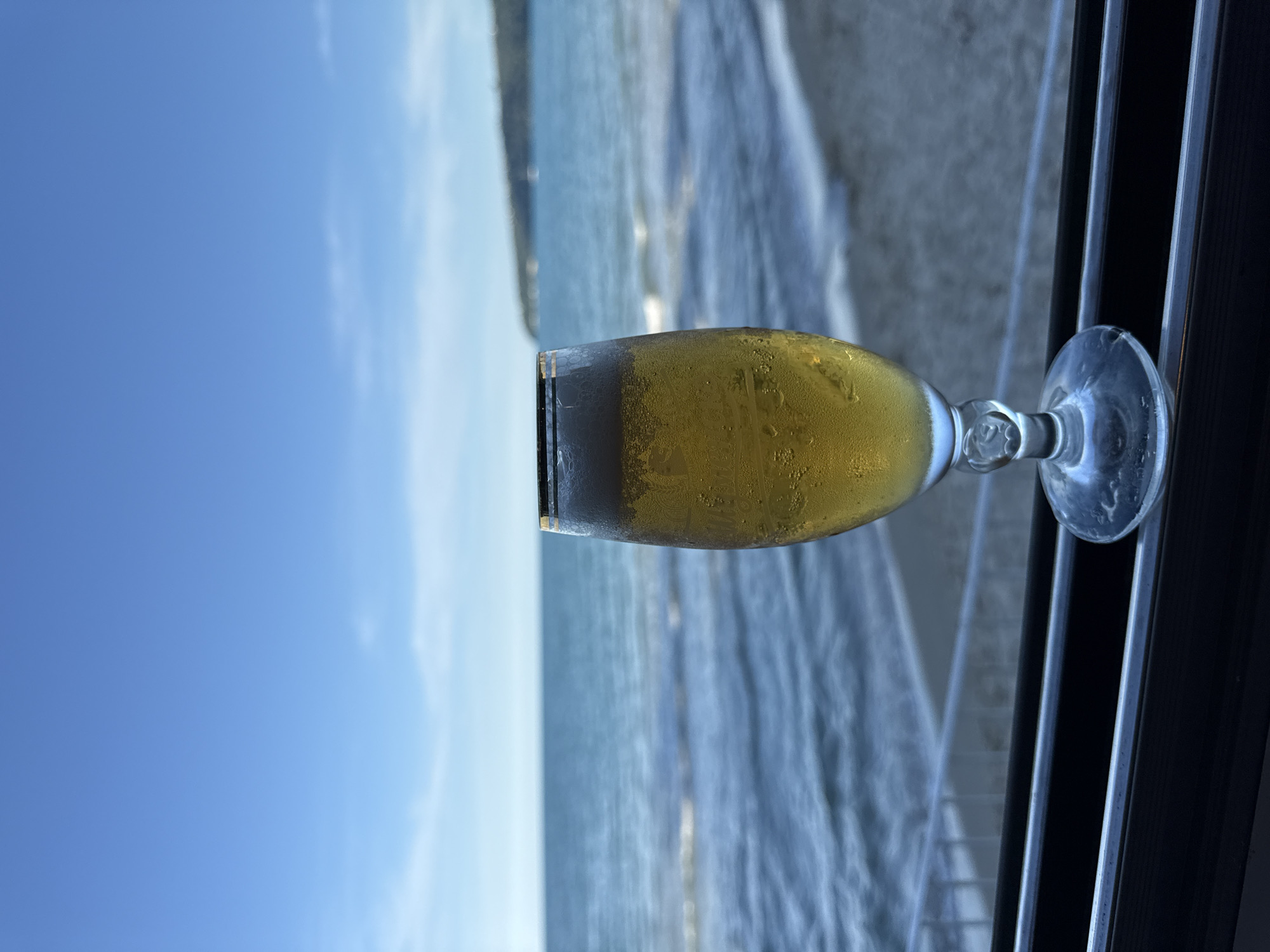 A clear glass of beer with a frothy head sits on a dark surface in the foreground, obscuring a railing. The beer is golden yellow and condensation beads on the outside of the glass. Beyond the glass, the ocean stretches out under a bright blue sky with wispy white clouds. Gentle waves lap at a sandy beach, and a distant, tree-covered headland is visible on the horizon. The scene appears to be captured from a seated perspective, perhaps at a restaurant or bar overlooking the water.