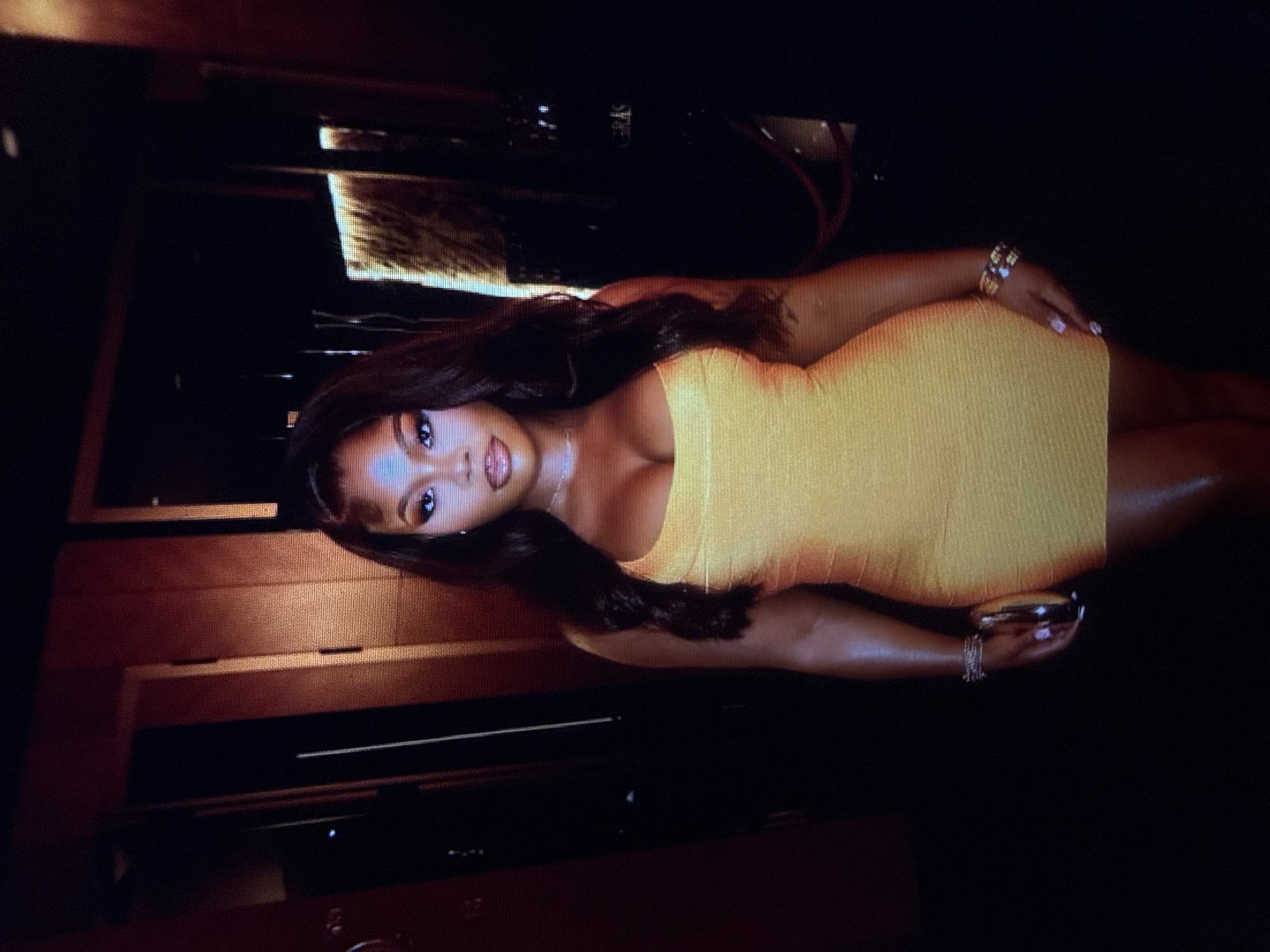 A woman with long, dark hair and a yellow bodycon dress stands in a dimly lit, indoor environment. She is wearing makeup and jewelry, including bracelets on both wrists and a necklace. She is holding a small, metallic clutch purse in her left hand. The background appears to be a hallway or lounge area with dark walls and some illuminated vertical elements. There is a red rope barrier visible in the right portion of the background. The image is a medium shot, focusing on the woman from the chest down. No specific location in Abuja, Nigeria can be determined from the visual information provided.