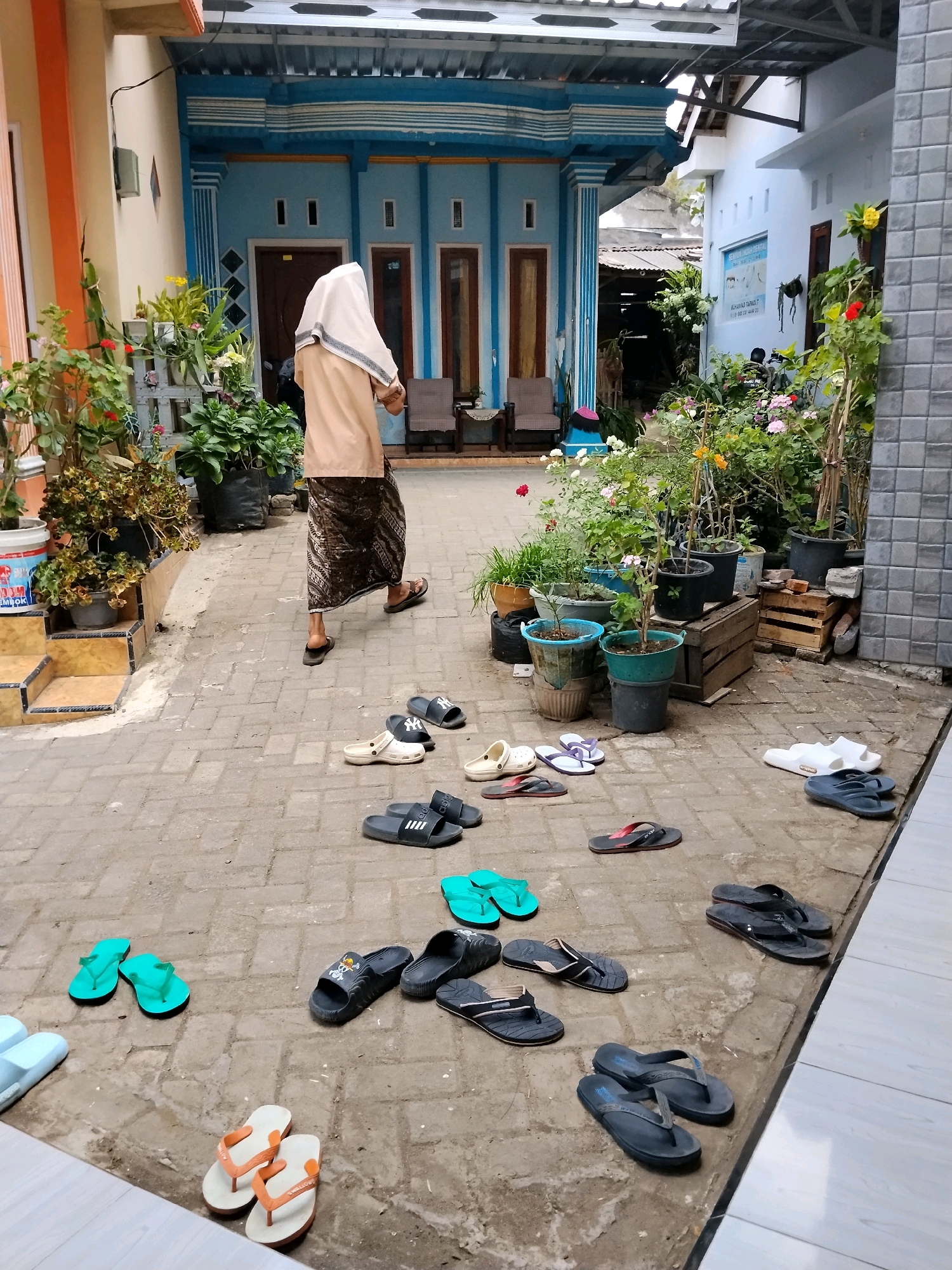 In Tlogosari, Indonesia, a person wearing a beige head covering and a batik sarong walks across a paved courtyard. The courtyard is lined with potted plants and flowers, and a building with blue and white accents is visible in the background. Various pairs of flip-flops and sandals are scattered on the ground, suggesting a communal living or religious space. The scene appears to be outdoors, with sunlight illuminating the area.