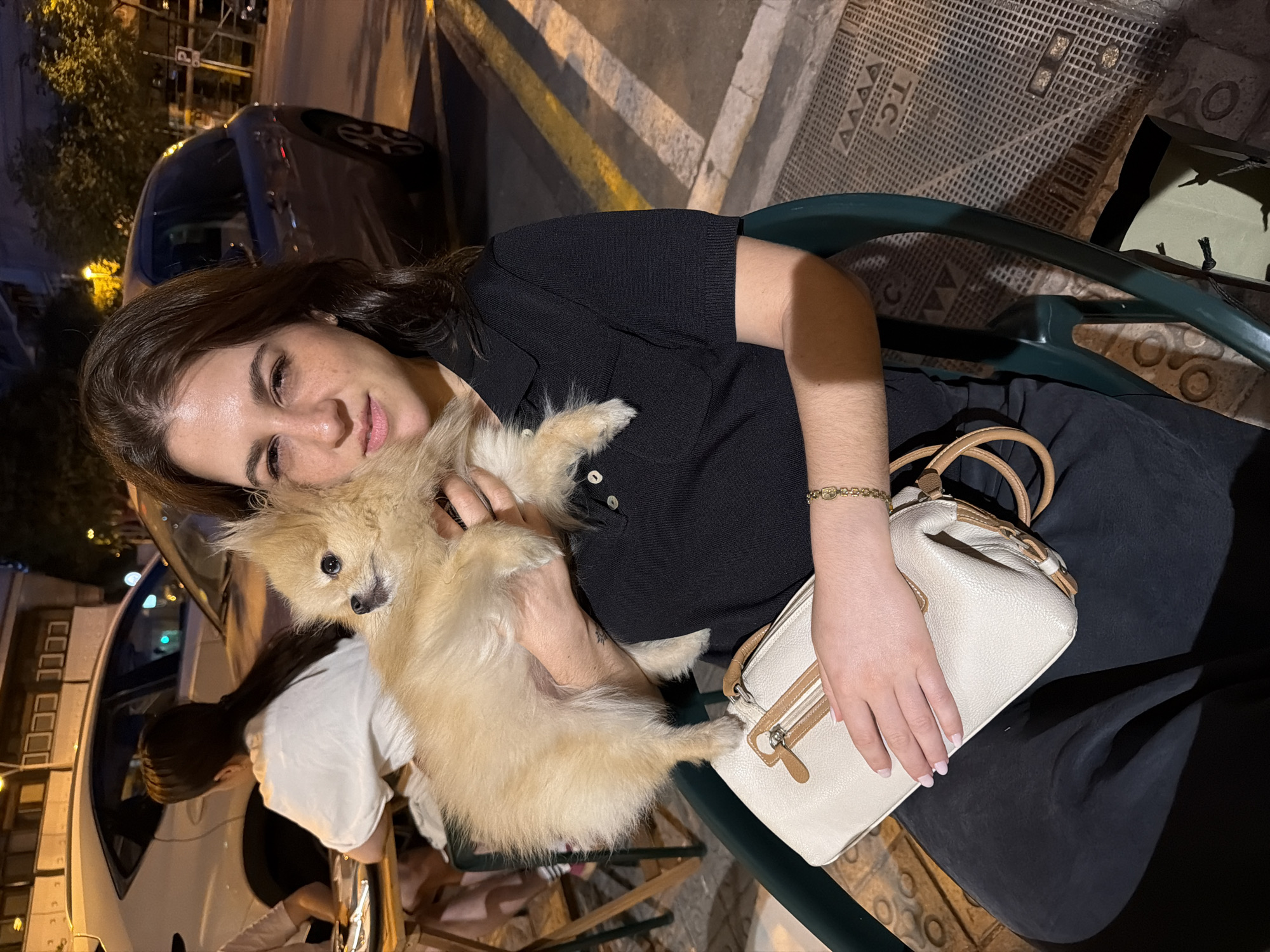 A woman is holding a small, fluffy dog and sitting on a dark green chair at an outdoor table. She is wearing a black collared shirt and dark pants. She has a light-colored purse on her lap. The setting appears to be a street at night, with cars and buildings visible in the background. The image was taken in Barcelona, Spain.