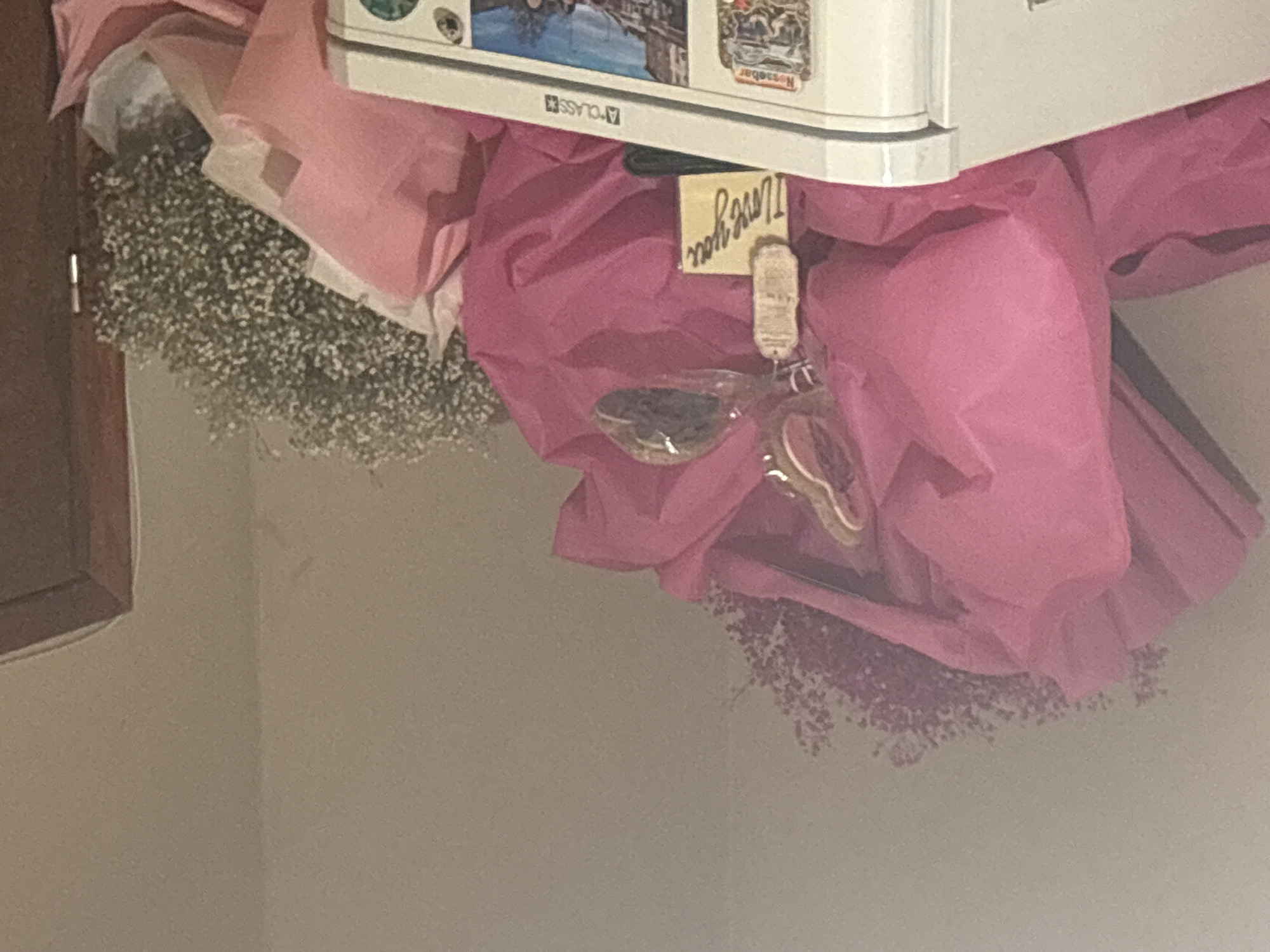 The image shows a gift arrangement. A bouquet of baby's breath flowers, wrapped in pink tissue paper, sits beneath what appears to be a small, white refrigerator. A small, handwritten "I love you" note is attached to the tissue paper, along with a clear glass container holding dark-colored contents and a decorative item. The scene is set against a plain, light-colored wall and a partially visible wooden door frame on the left side of the image. The location appears to be indoors.