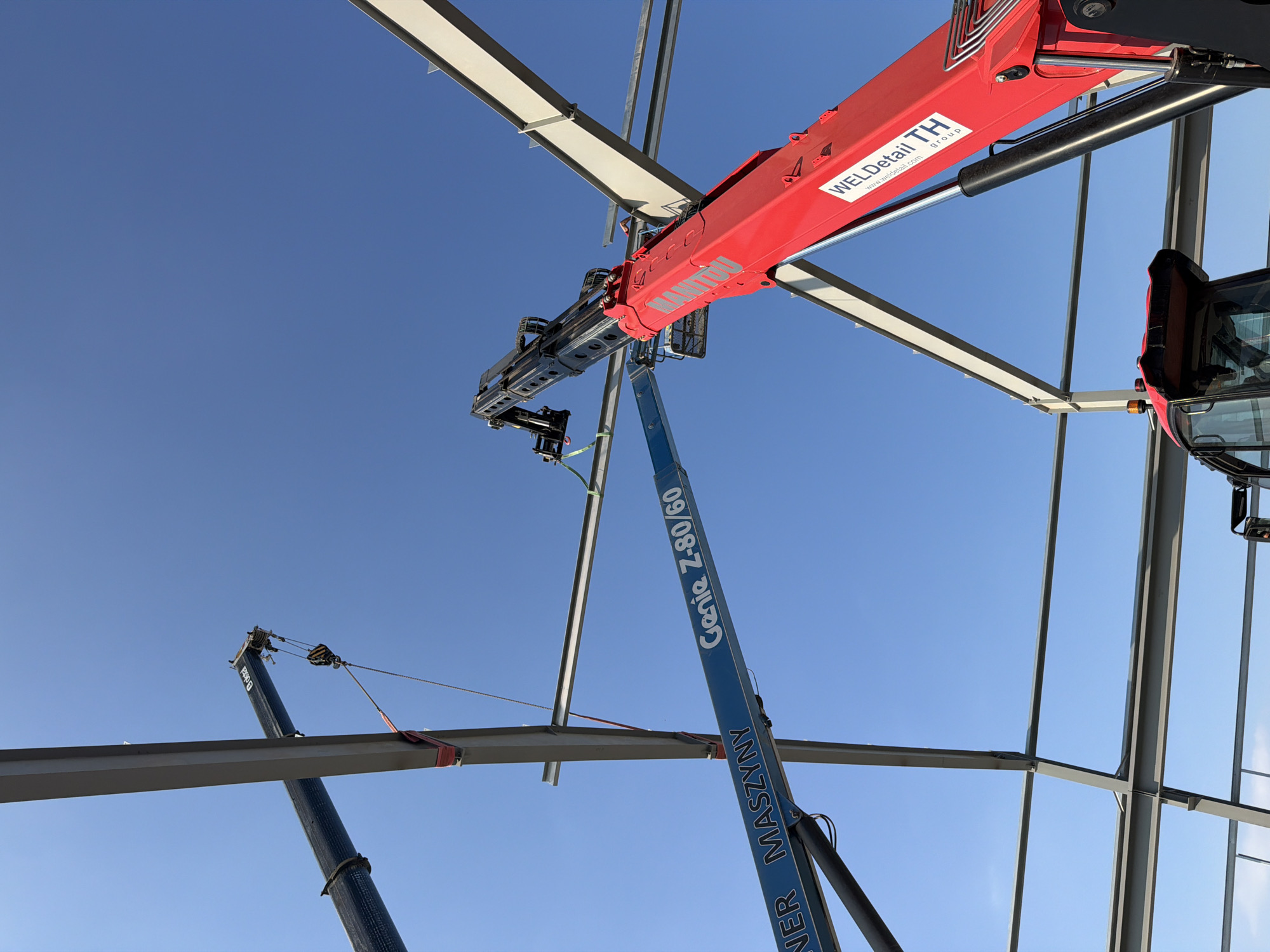The image captures an outdoor construction scene under a clear blue sky in Stróża, Poland. The primary focus is on the assembly of a steel structure, with large gray I-beams forming the framework of a building. Two cranes are actively engaged in lifting and positioning these beams.
On the left, a black crane arm extends upwards, holding a steel beam suspended by red and green straps. A yellow and black hook mechanism is visible at the end of the crane's cable. Adjacent to this crane, a blue boom lift is positioned, displaying the text "NER MASZYNY Genie Z-80/60".
To the right, a large red telescopic crane, identified by the "MANITOU" logo and "WELDdetail TH group" branding, is prominently featured. Its extended boom is angled upwards, reaching towards the steel beams. A portion of a red construction vehicle, likely the base of the Manitou crane, is visible at the bottom of the frame, showing a black cab with a yellow flashing light. The steel beams are connected at various points, indicating ongoing construction progress. The ground is not visible in the image, but the overall impression is one of active construction work in an open area.